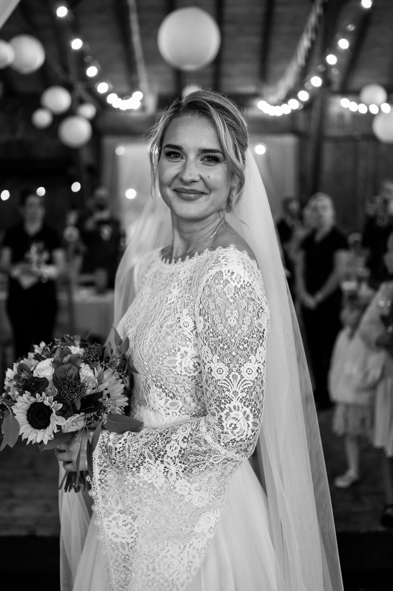 Portret uśmiechniętej panny młodej w koronkowej sukni z bukietem słoneczników, welonem i delikatną biżuterią na tle sali z dekoracyjnym oświetleniem.