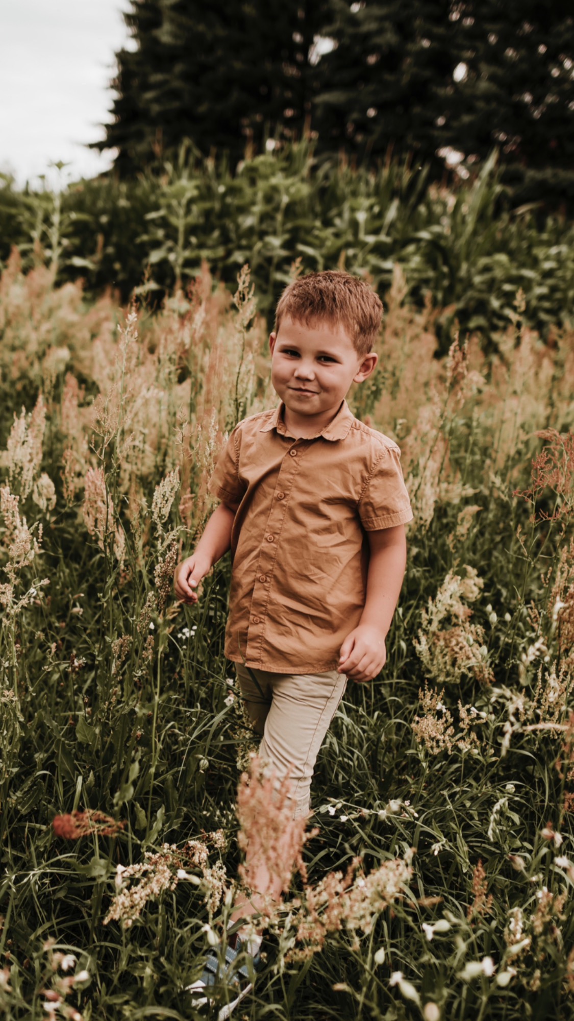 Uśmiechnięty chłopiec w brązowej koszuli i beżowych spodniach stoi na łące wśród wysokich traw i kwiatów, patrząc w obiektyw.