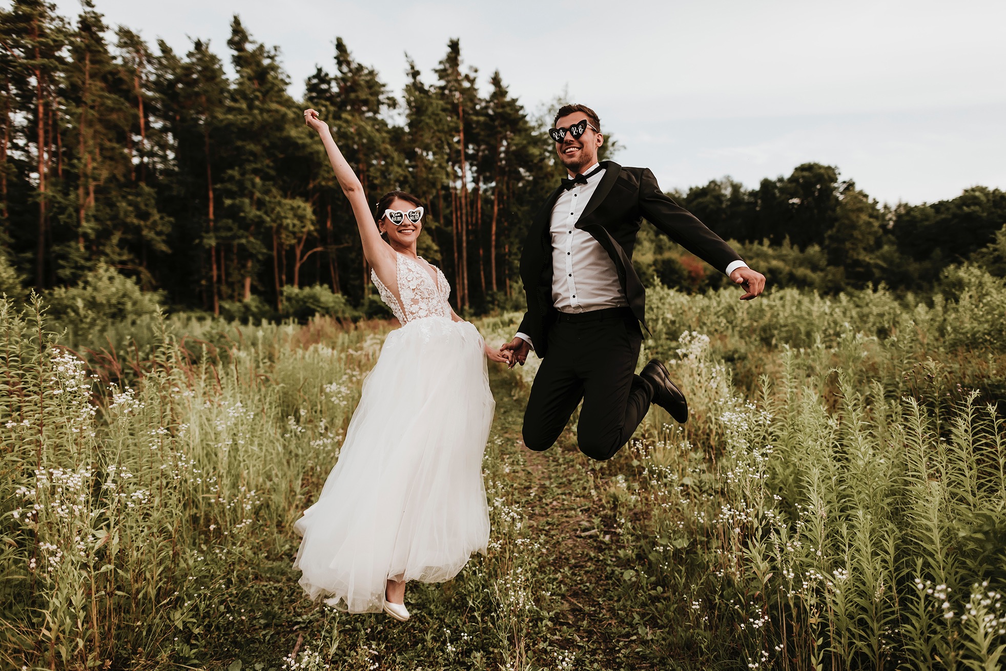 Uśmiechnięta para młoda w skoku na tle łąki i lasu, oboje w okularach w kształcie serc z napisem 'bride' i 'groom'.