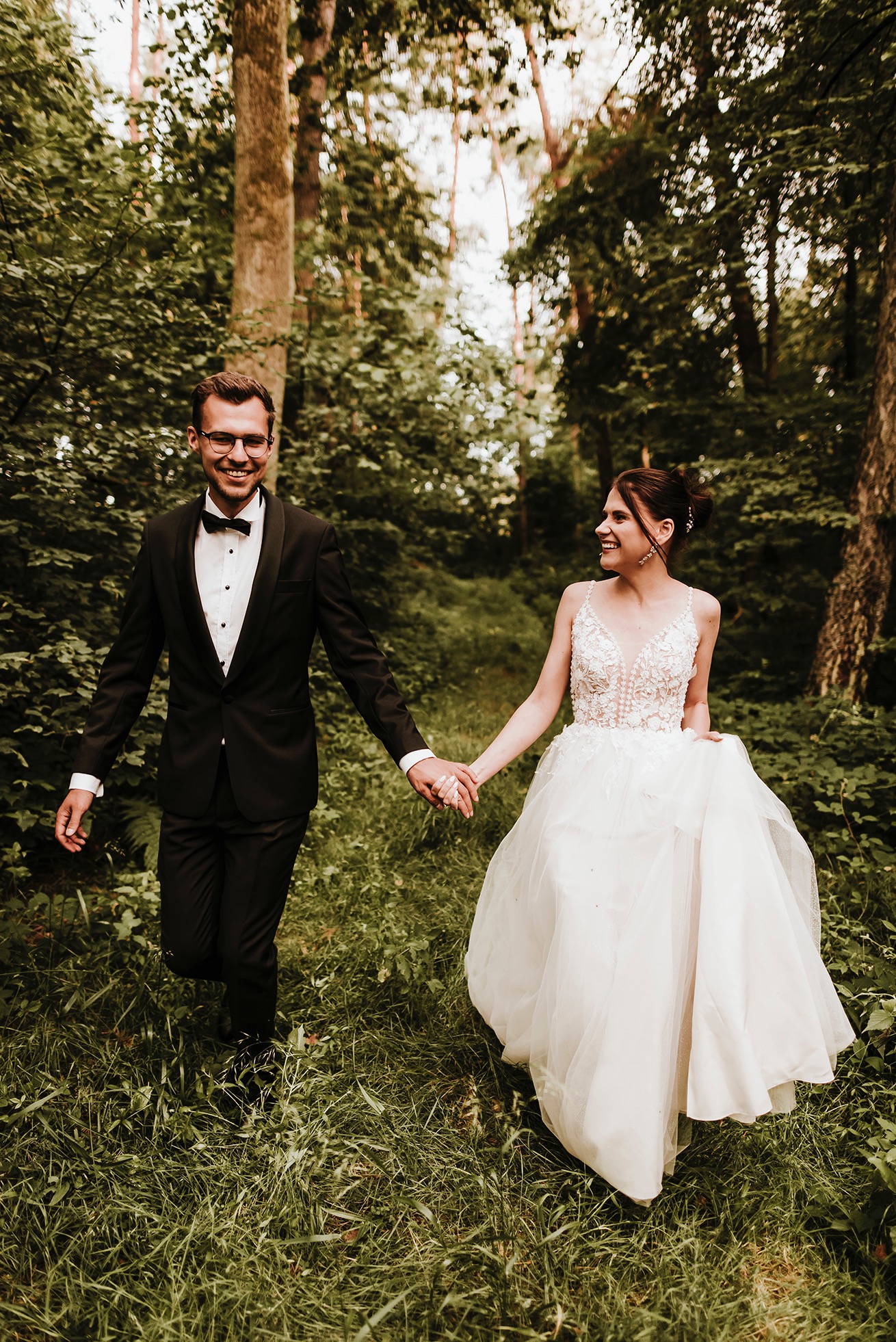 Uśmiechnięta para młoda, on w czarnym smokingu i okularach, ona w białej sukni z koronkową górą, trzymają się za ręce idąc przez zarośnięty las.