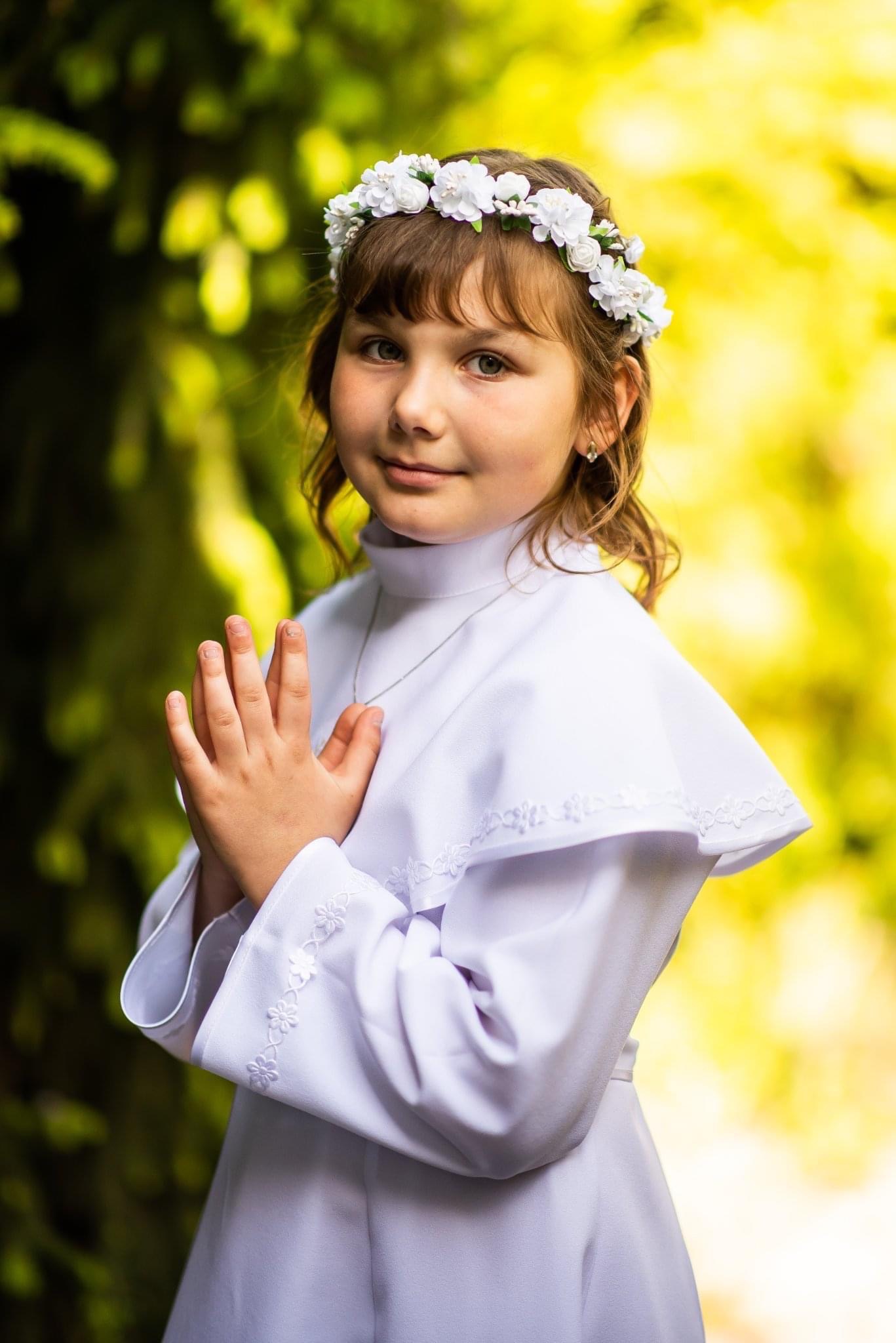 Portret dziewczynki w białej albie komunijnej z wiankiem z białych kwiatów na głowie, złożone dłonie, delikatny uśmiech, na tle zieleni.