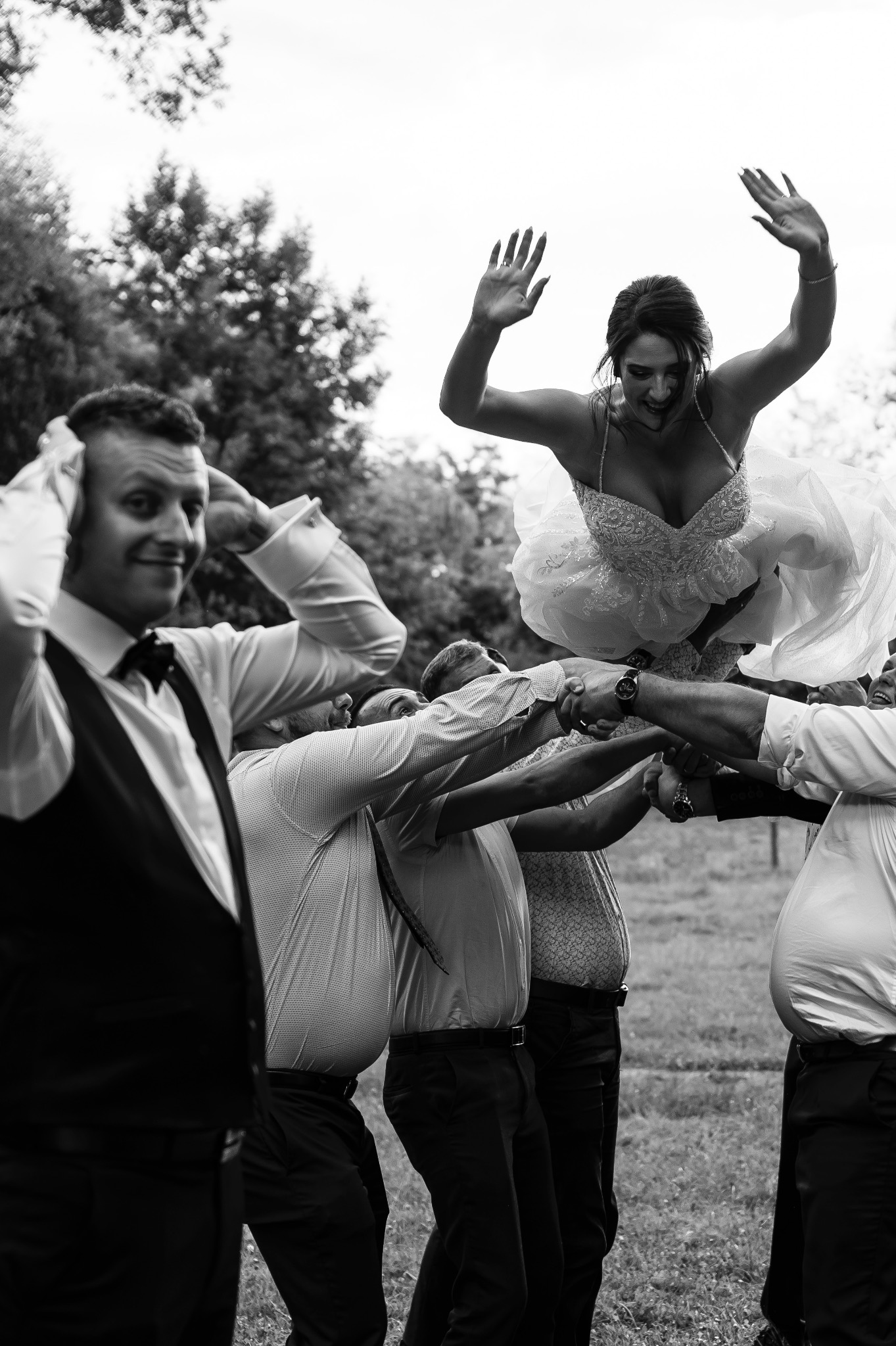 Czarno-białe zdjęcie panny młodej unoszonej przez grupę mężczyzn, z uśmiechem i uniesionymi rękami na tle drzew i nieba.