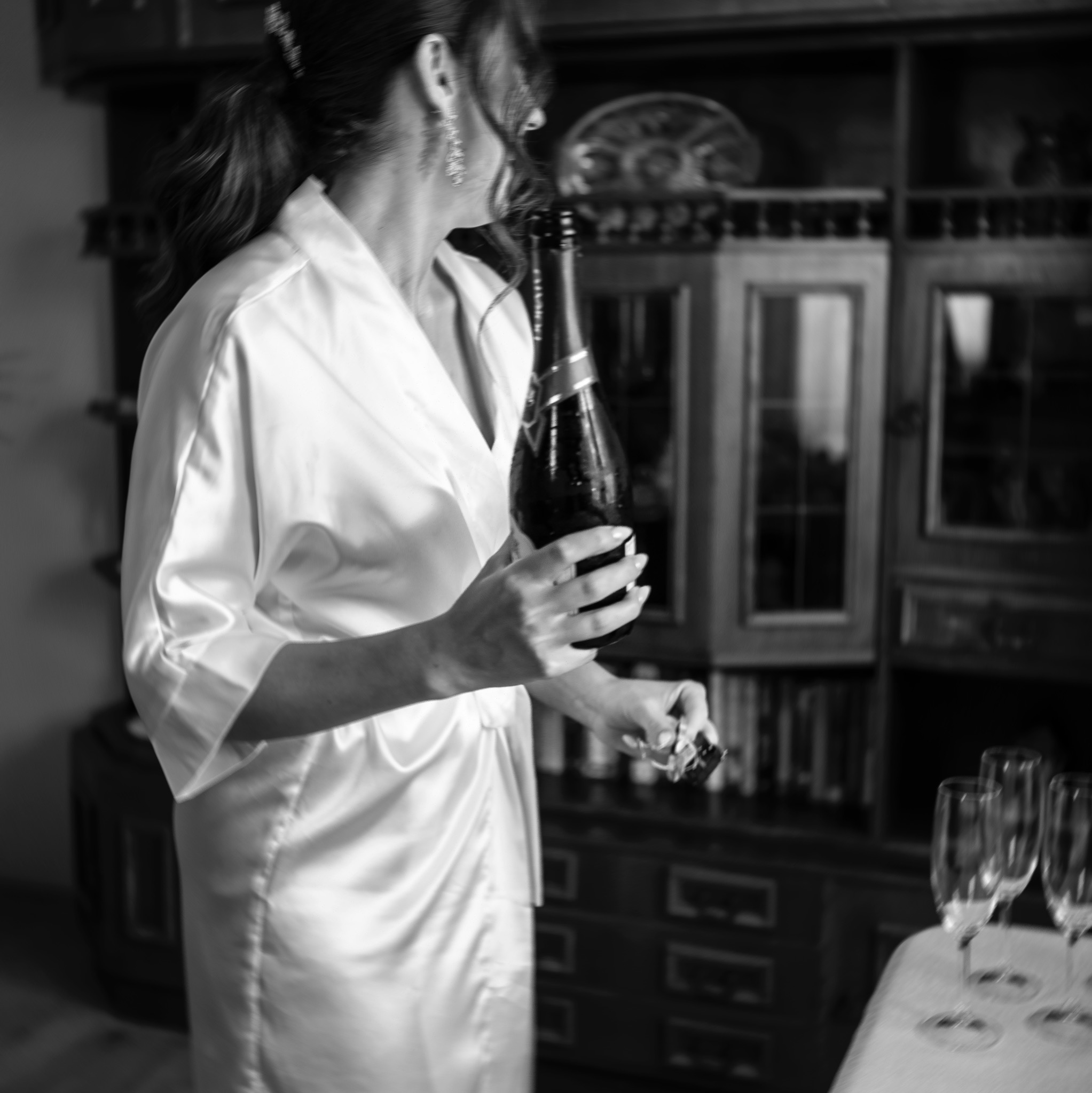 Panna młoda w satynowym szlafroku otwiera butelkę szampana przed ślubem, w tle kredens i kieliszki.