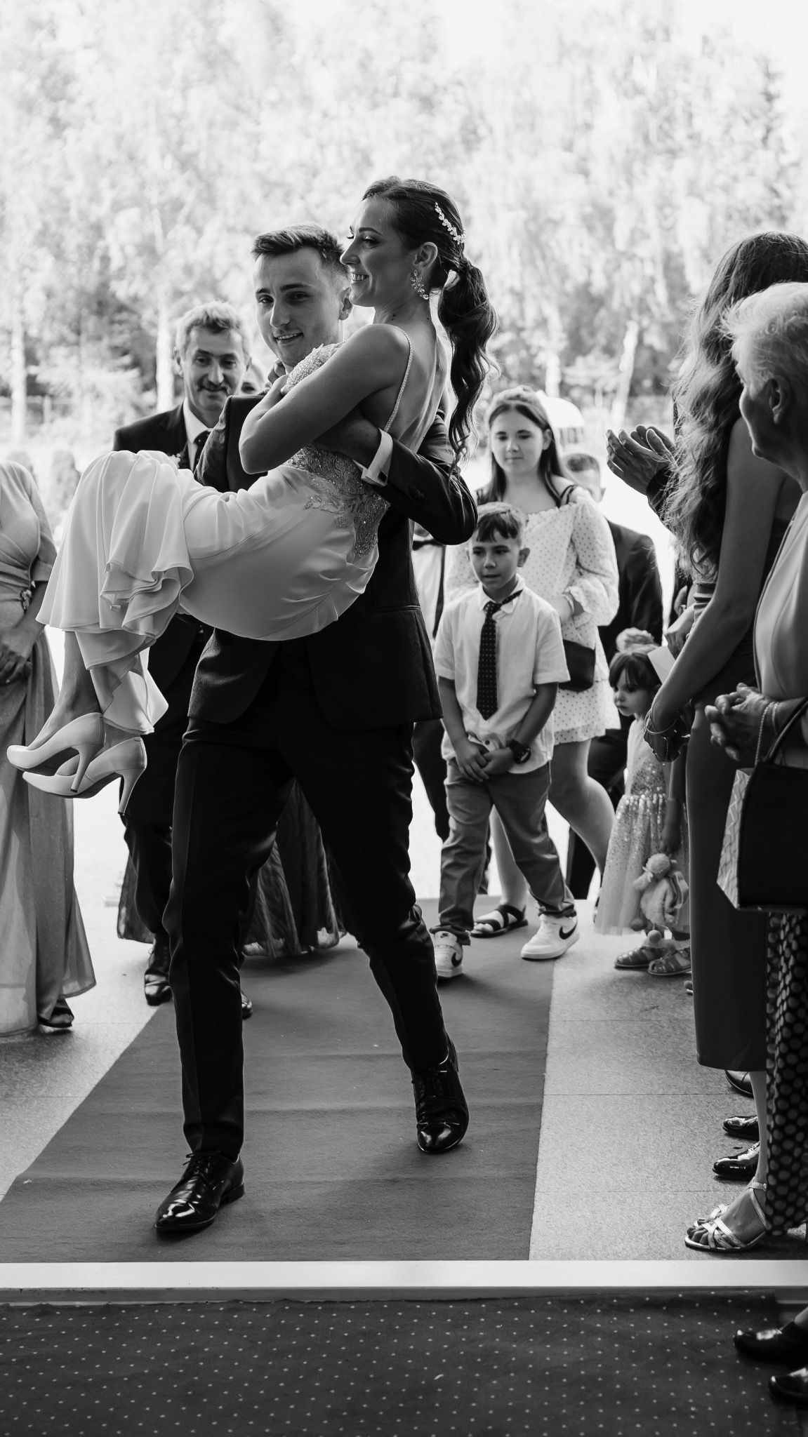 Pan młody niesie uśmiechniętą pannę młodą na rękach po czerwonym dywanie, w tle goście weselni i dziecko w krawacie.