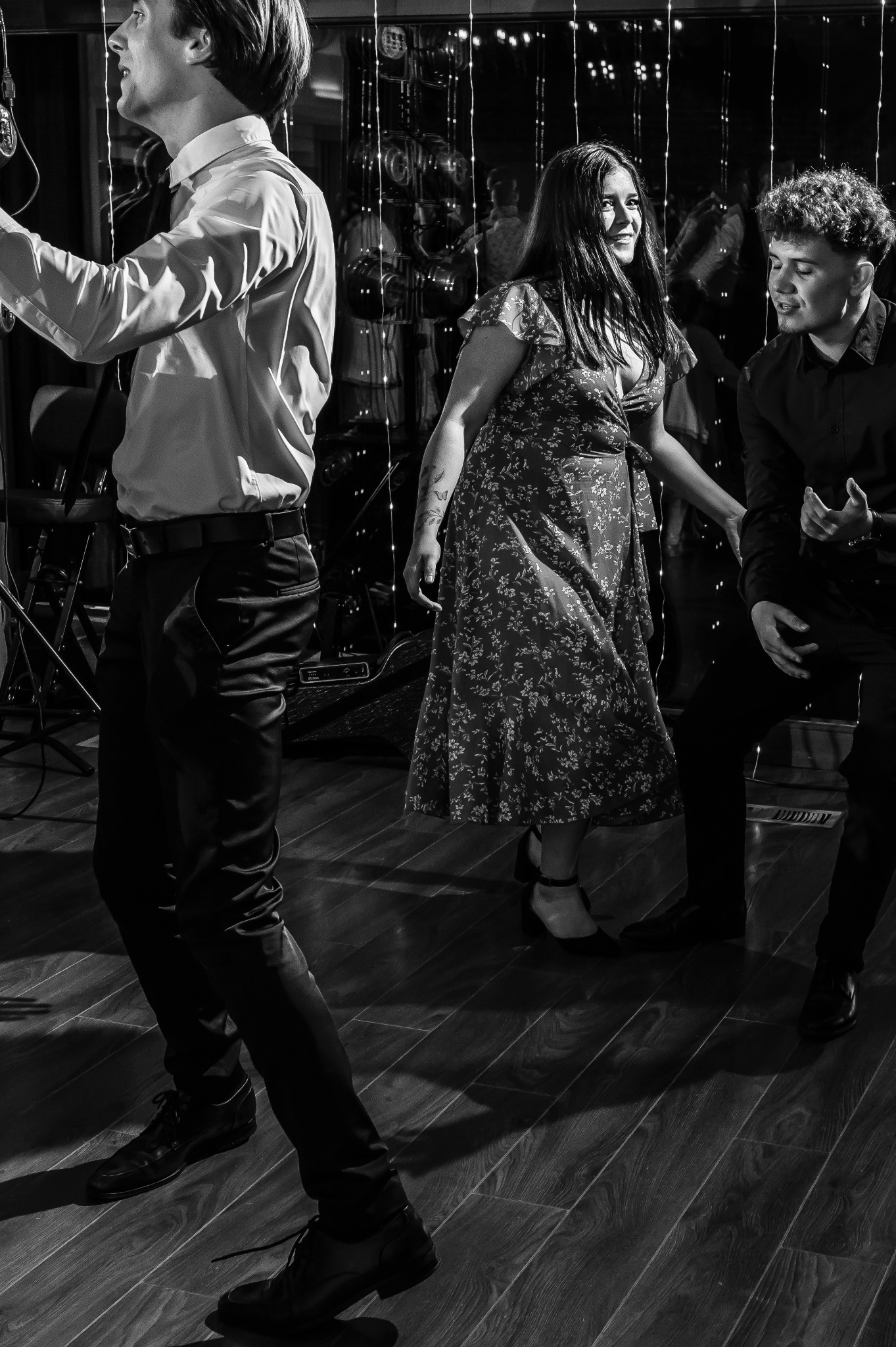 Czarno-białe zdjęcie przedstawia tańczących ludzi na parkiecie, oświetlonym przez dekoracyjne lampki. Kobieta w kwiecistej sukience uśmiecha się, obok niej mężczyzna w czarnej koszuli. W tle...