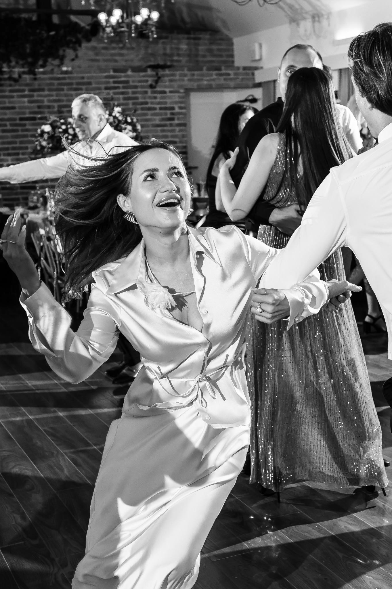 Czarno-białe zdjęcie rozbawionej kobiety w satynowej sukience, tańczącej na parkiecie z innymi gośćmi podczas wydarzenia, z ceglaną ścianą w tle i widocznym oświetleniem.