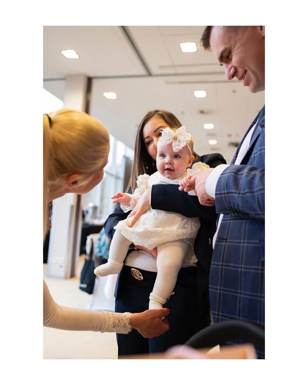 Uśmiechnięta niemowlę w białej sukience z koronkowymi wykończeniami i dużą kokardą na głowie, trzymana na rękach przez mężczyznę w granatowym garniturze w kratę, otoczona przez dwie kobiety,...