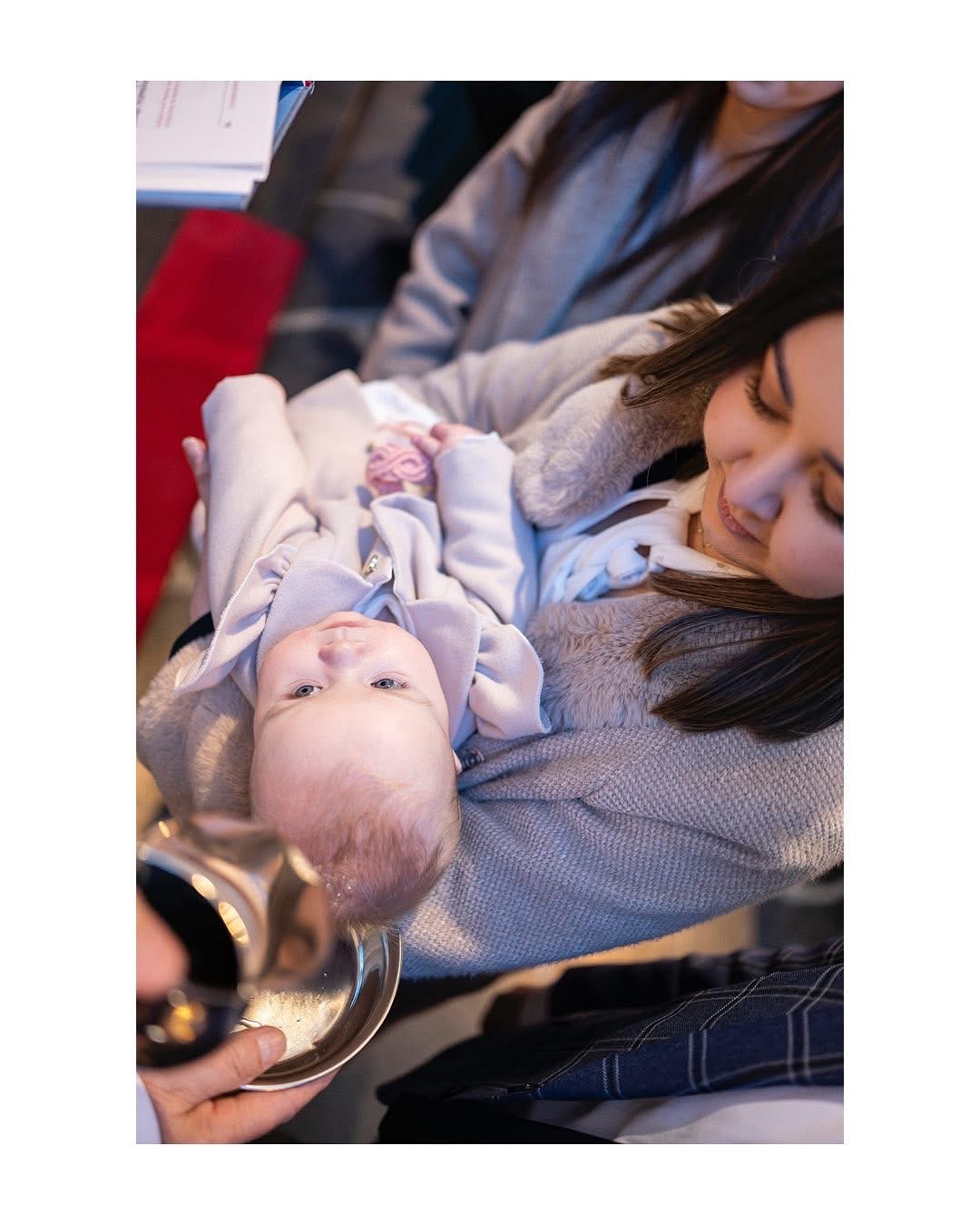 Kąpiel niemowlęcia podczas ceremonii, w tle uśmiechnięta kobieta z ciemnymi włosami.