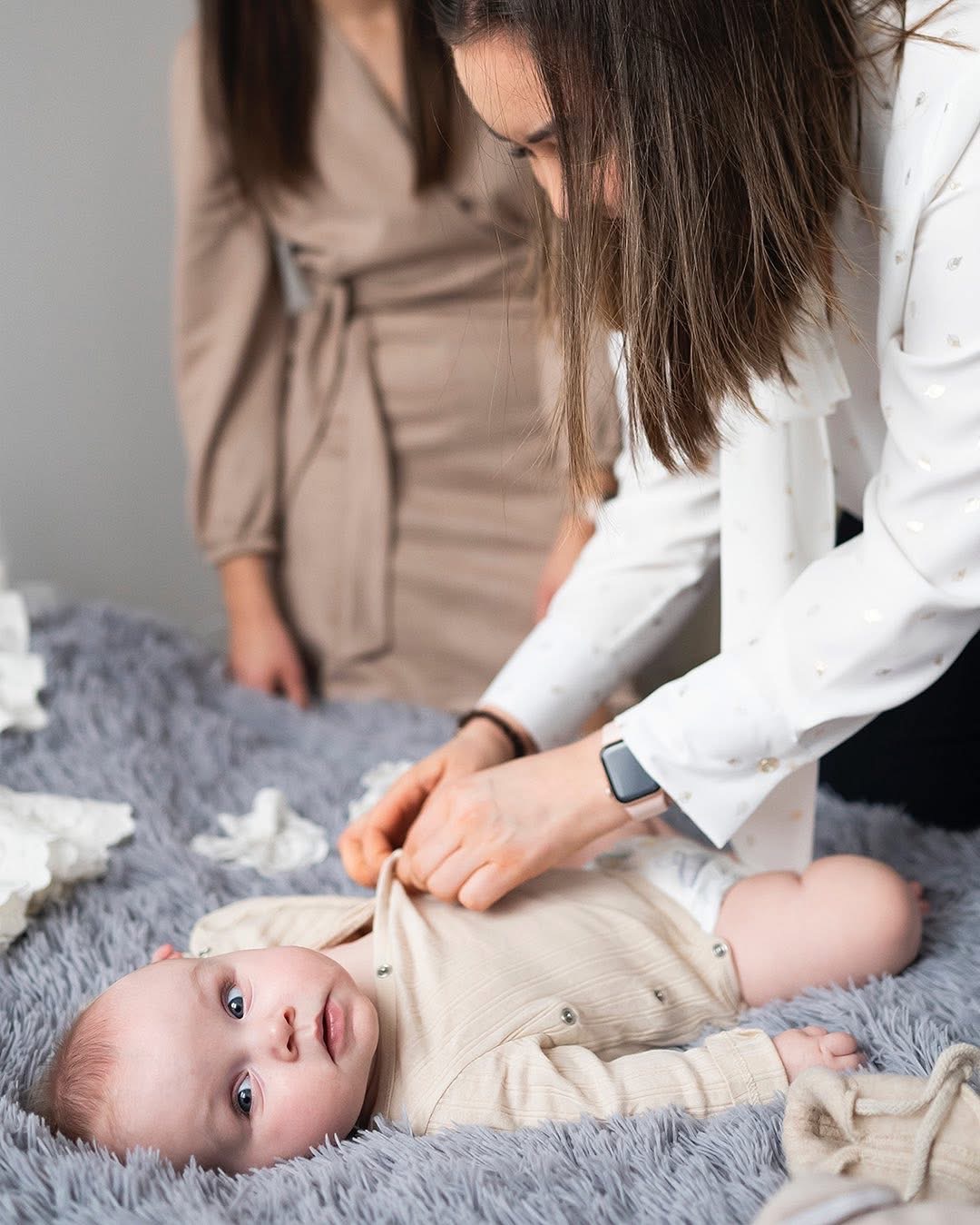 Niemowlę leżące na puszystym szarym kocyku, ubrane w beżowe body, podczas ubierania przez kobietę w białej koszuli z delikatnym wzorem, w tle druga kobieta w beżowej sukience.