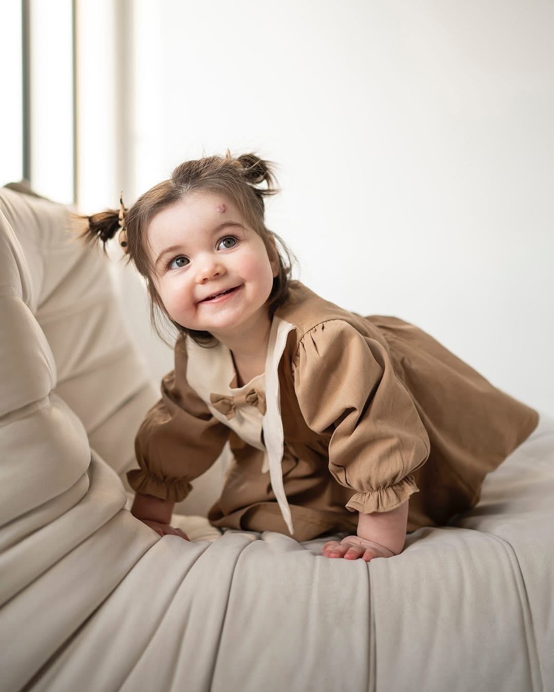 Uśmiechnięta dziewczynka w brązowej sukience z kokardą, raczkuje na jasnej sofie, z dwoma kucykami i małą ranką na czole.