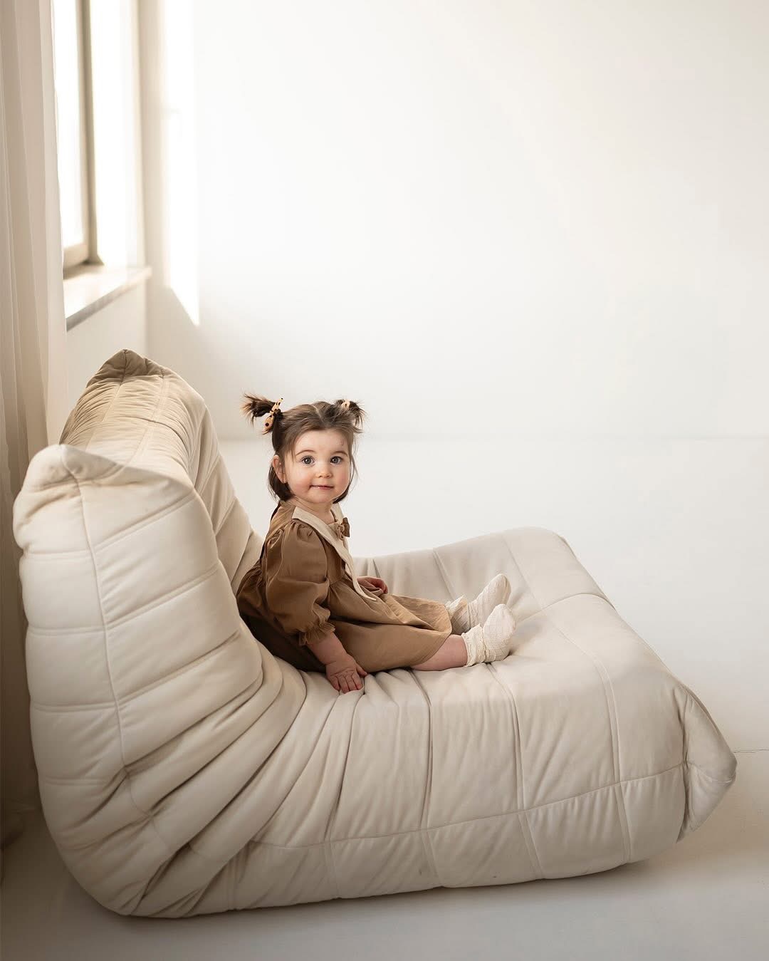 Uśmiechnięta dziewczynka z brązowymi włosami spiętymi w dwa kucyki siedzi na kremowym, modułowym fotelu w jasnym, minimalistycznym wnętrzu.