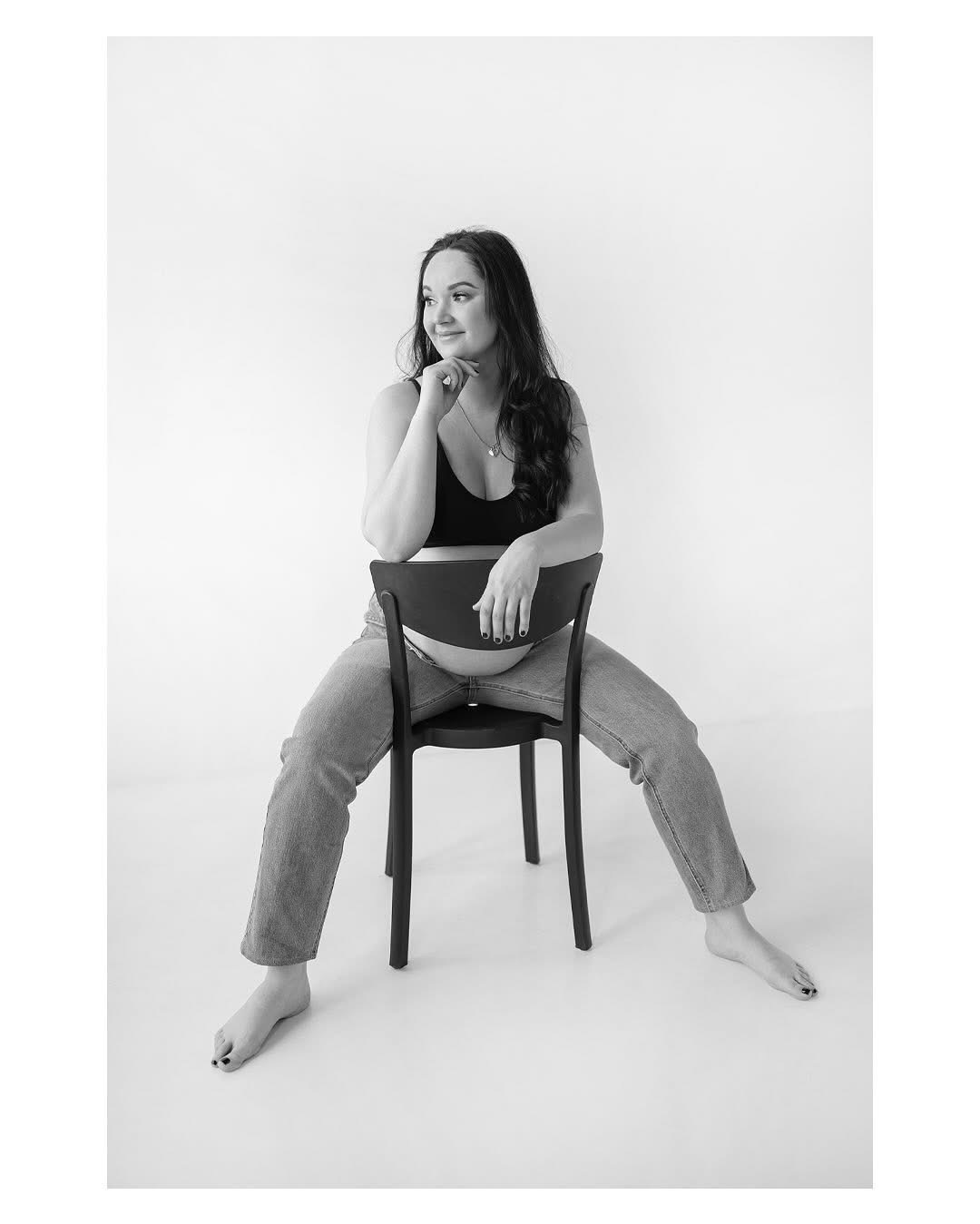 Czarno-białe zdjęcie kobiety w ciąży siedzącej tyłem na krześle, w dżinsach i topie, z ręką podpartą pod brodą, zamyślony wzrok skierowany w lewo.