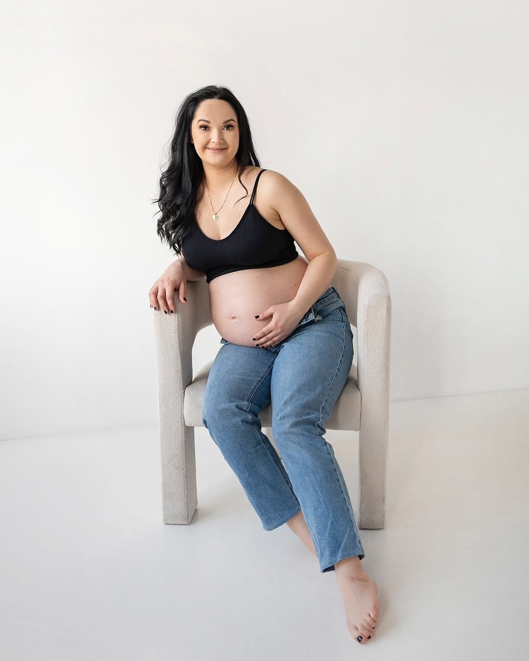 Kobieta w ciąży, w czarnym topie i jeansach, siedzi na krześle na białym tle, trzymając ręce na brzuchu i uśmiechając się.