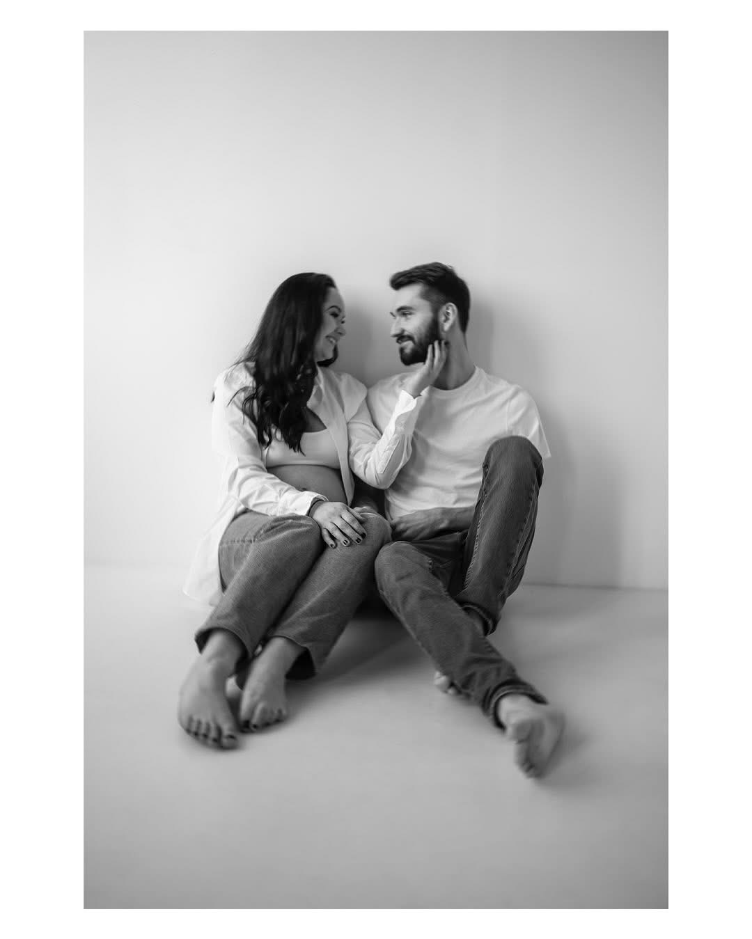 Czarno-białe zdjęcie pary siedzącej na podłodze: kobieta w ciąży w rozpiętej białej koszuli i jeansach, mężczyzna z brodą w białym t-shircie i jeansach, delikatnie dotyka jej twarzy, oboje patrzą...