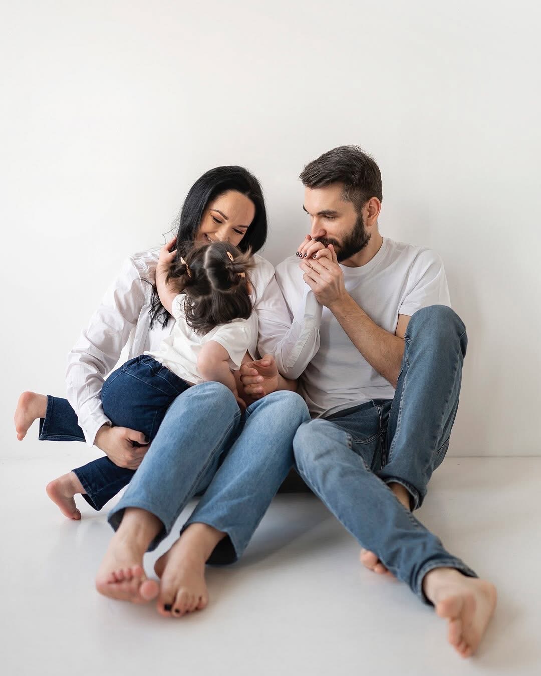 Rodzina siedząca na podłodze: mama trzyma córkę na kolanach, tata całuje dłoń córki, wszyscy ubrani w dżinsy i białe koszulki, minimalistyczne tło.