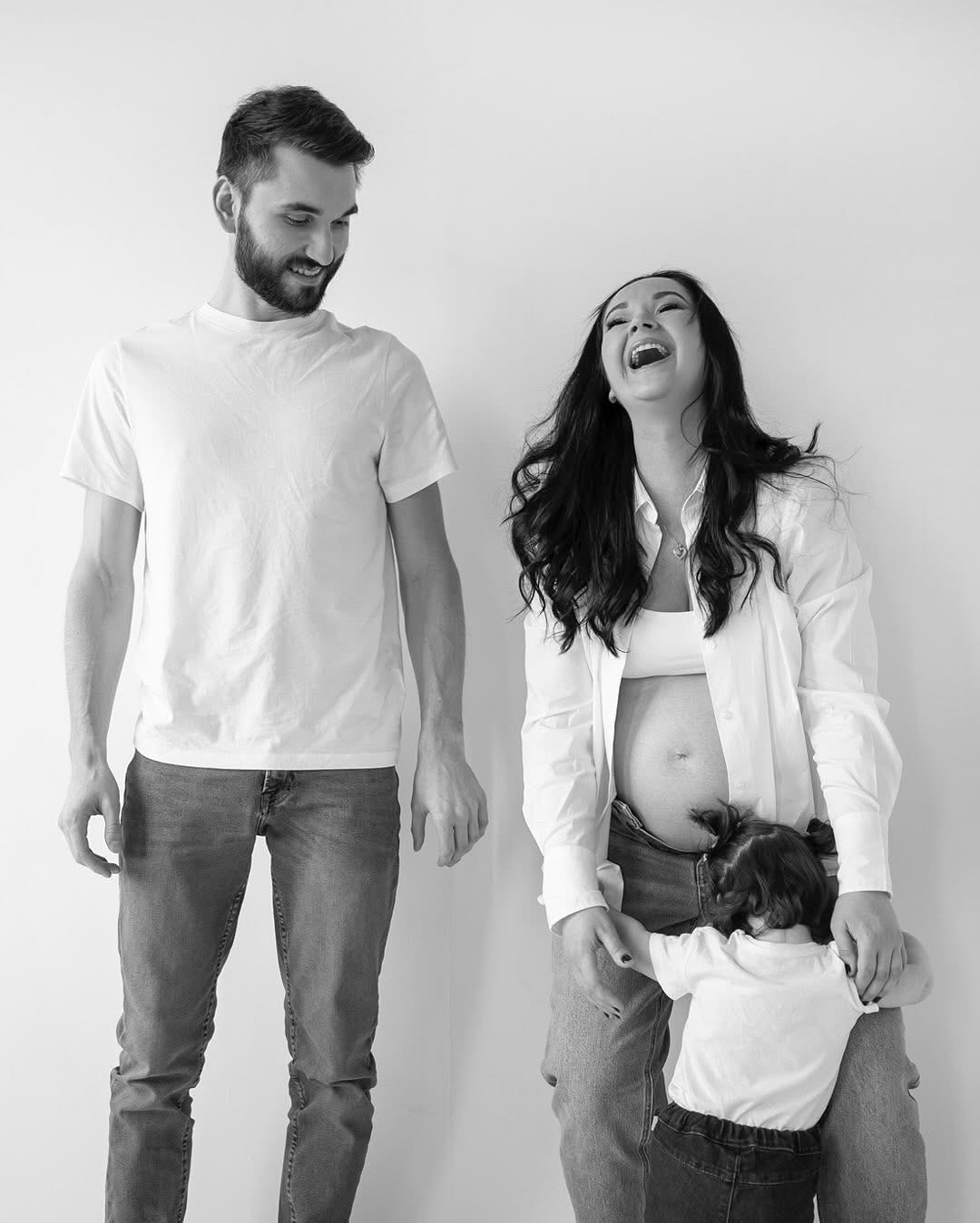 Czarno-białe zdjęcie szczęśliwej rodziny w Niepołomicach: uśmiechnięty ojciec, śmiejąca się matka w ciąży i mała dziewczynka przytulająca się do jej brzucha.