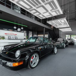 Eradur Sp. z o.o. - Salon samochodowy z czarnym Porsche 911 na pierwszym planie, szara posadzka z czerwoną linią, nowoczesne oświetlenie i recepcja w tle. Wnętrze Performance Center.