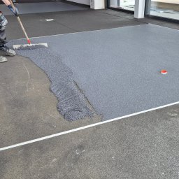 Eradur Sp. z o.o. - Aplikacja szarej posadzki żywicznej na podłożu. Widoczne narzędzie pracy i częściowo ukończona powierzchnia. W tle okno i materiały budowlane.