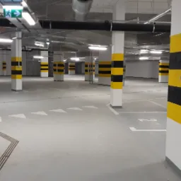Szare posadzki w garażu podziemnym z białymi słupami pomalowanymi w czarno-żółte pasy ostrzegawcze, widoczne oznaczenia poziome i system wentylacji pod sufitem.