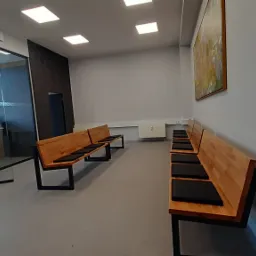 Jasnoszara posadzka w poczekalni z drewnianymi ławkami z czarnymi poduszkami, widok na przeszklone biuro i obraz na ścianie.