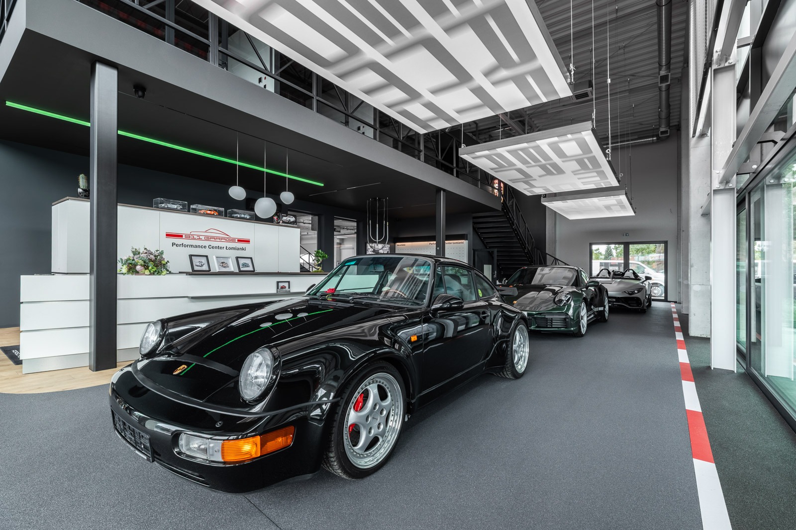 Salon samochodowy z czarnym Porsche 911 na pierwszym planie, szara posadzka z czerwoną linią, nowoczesne oświetlenie i recepcja w tle. Wnętrze Performance Center.