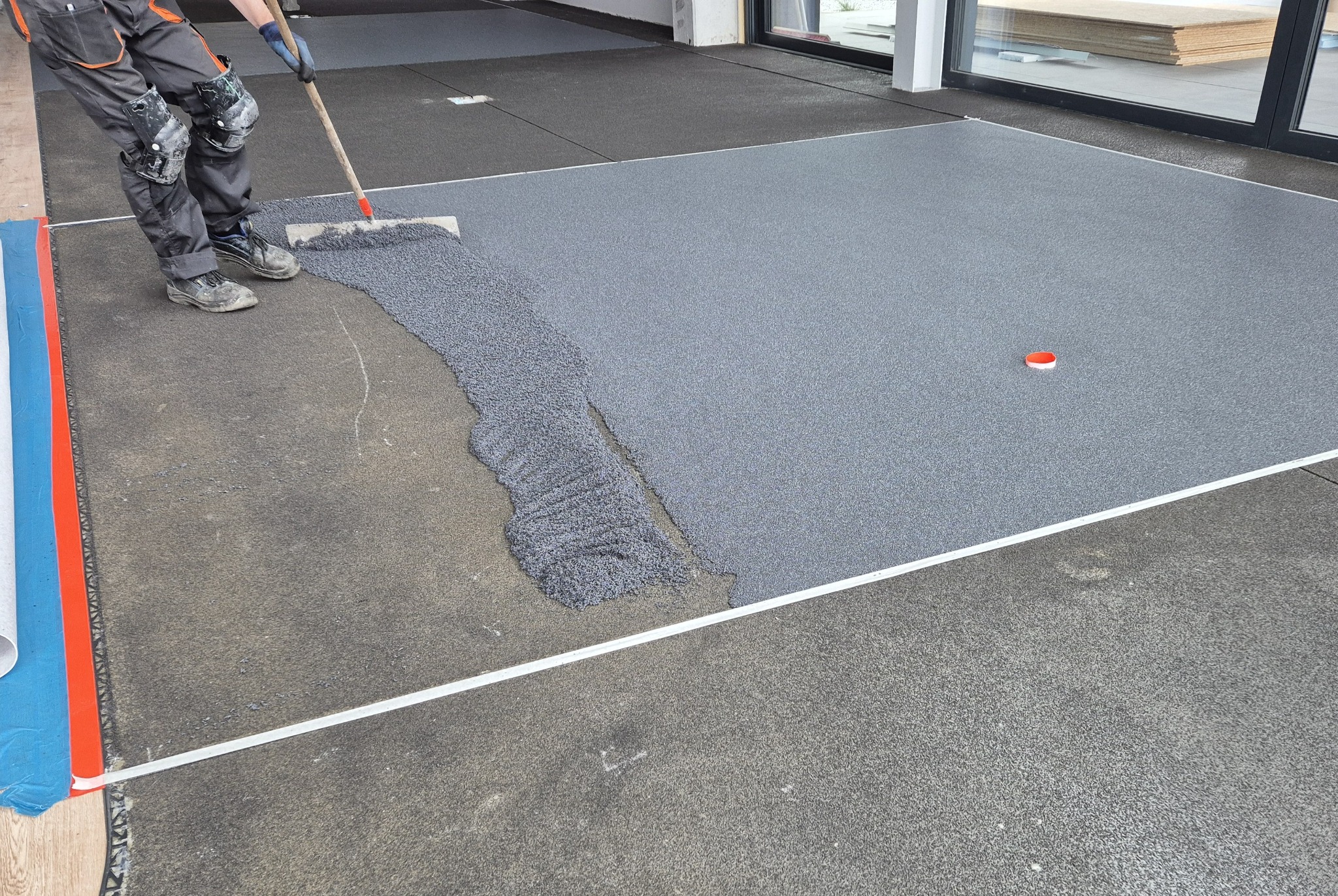 Aplikacja szarej posadzki żywicznej na podłożu. Widoczne narzędzie pracy i częściowo ukończona powierzchnia. W tle okno i materiały budowlane.