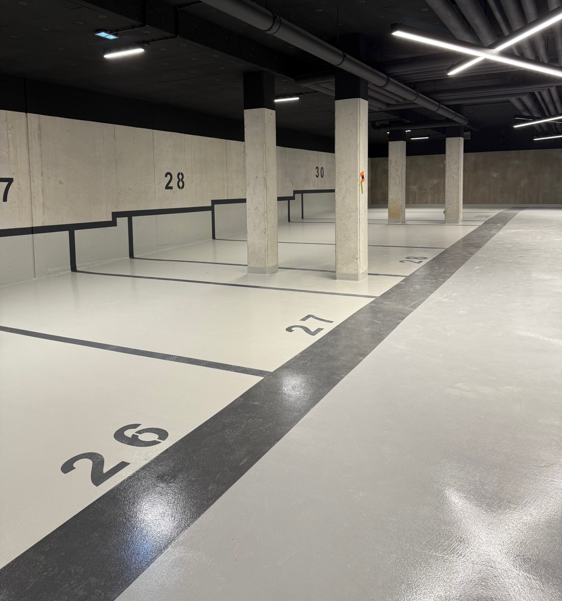 Jasna posadzka żywiczna w garażu podziemnym z numeracją miejsc parkingowych na ścianach i podłodze. Widoczne betonowe filary i czarne rury pod sufitem.