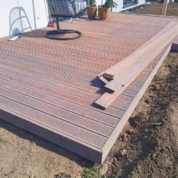 Taras z deski kompozytowej w trakcie budowy, widoczne deski ułożone na części powierzchni, obok leżą niezamontowane elementy, w tle dom z białym tynkiem i wiszący fotel.