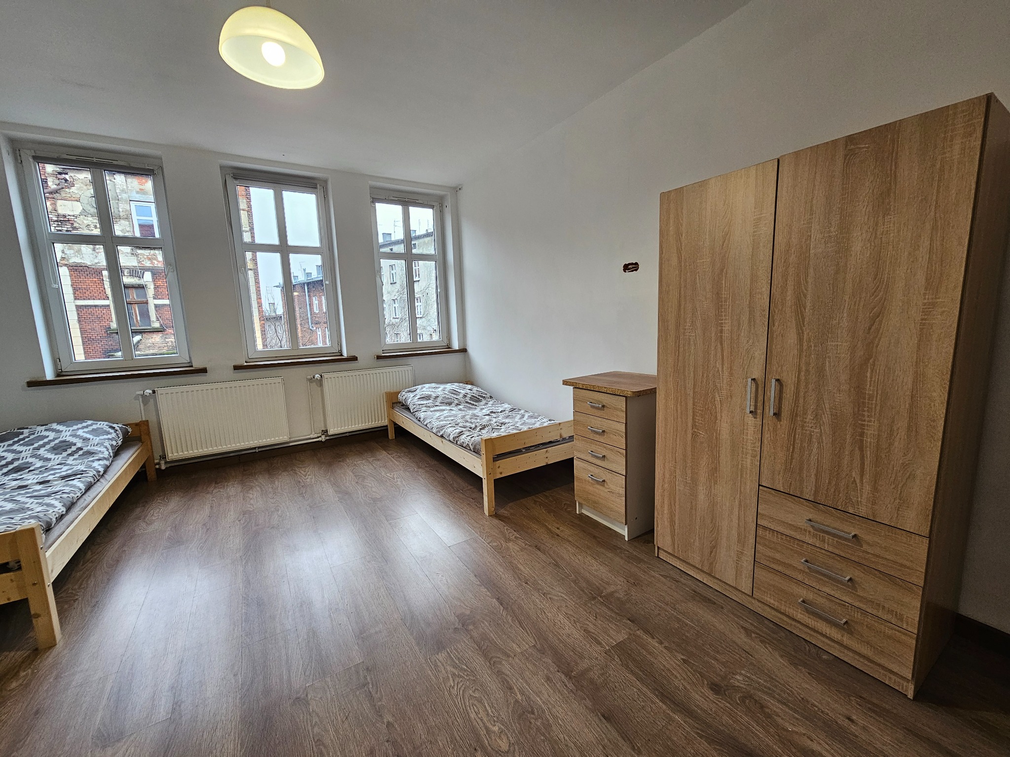Pokój z dwoma łóżkami pojedynczymi, drewnianą szafą, komodą i parkietem, widok z okna na budynki z cegły.