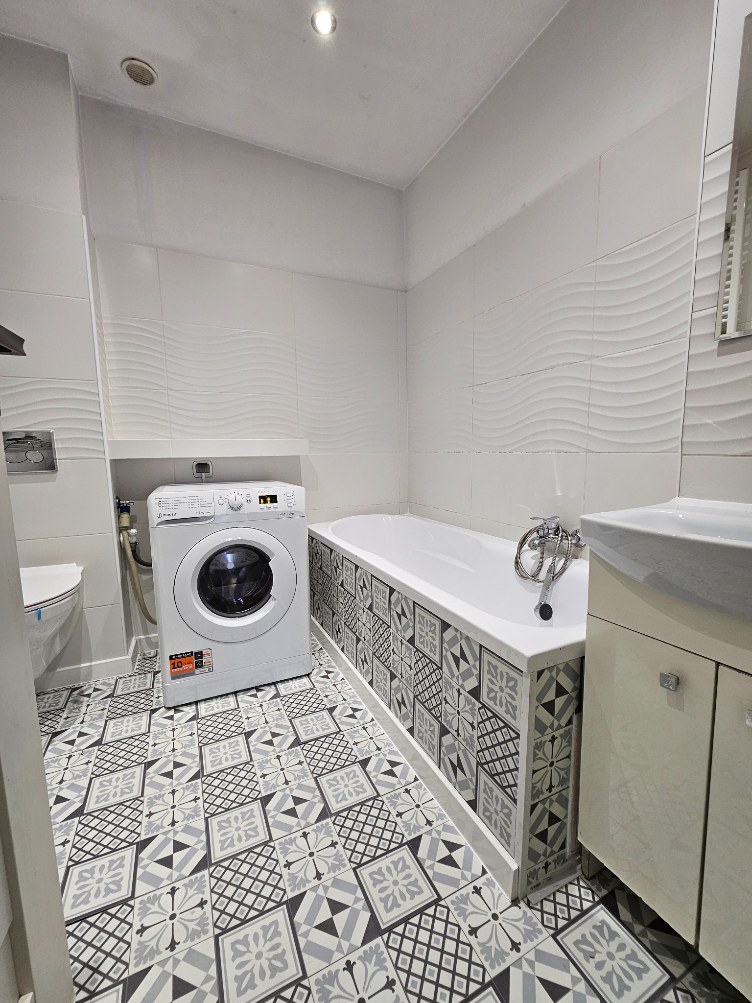 Nowoczesna łazienka z białą pralką Indesit, wzorzystą podłogą i obudową wanny w geometryczne wzory.
