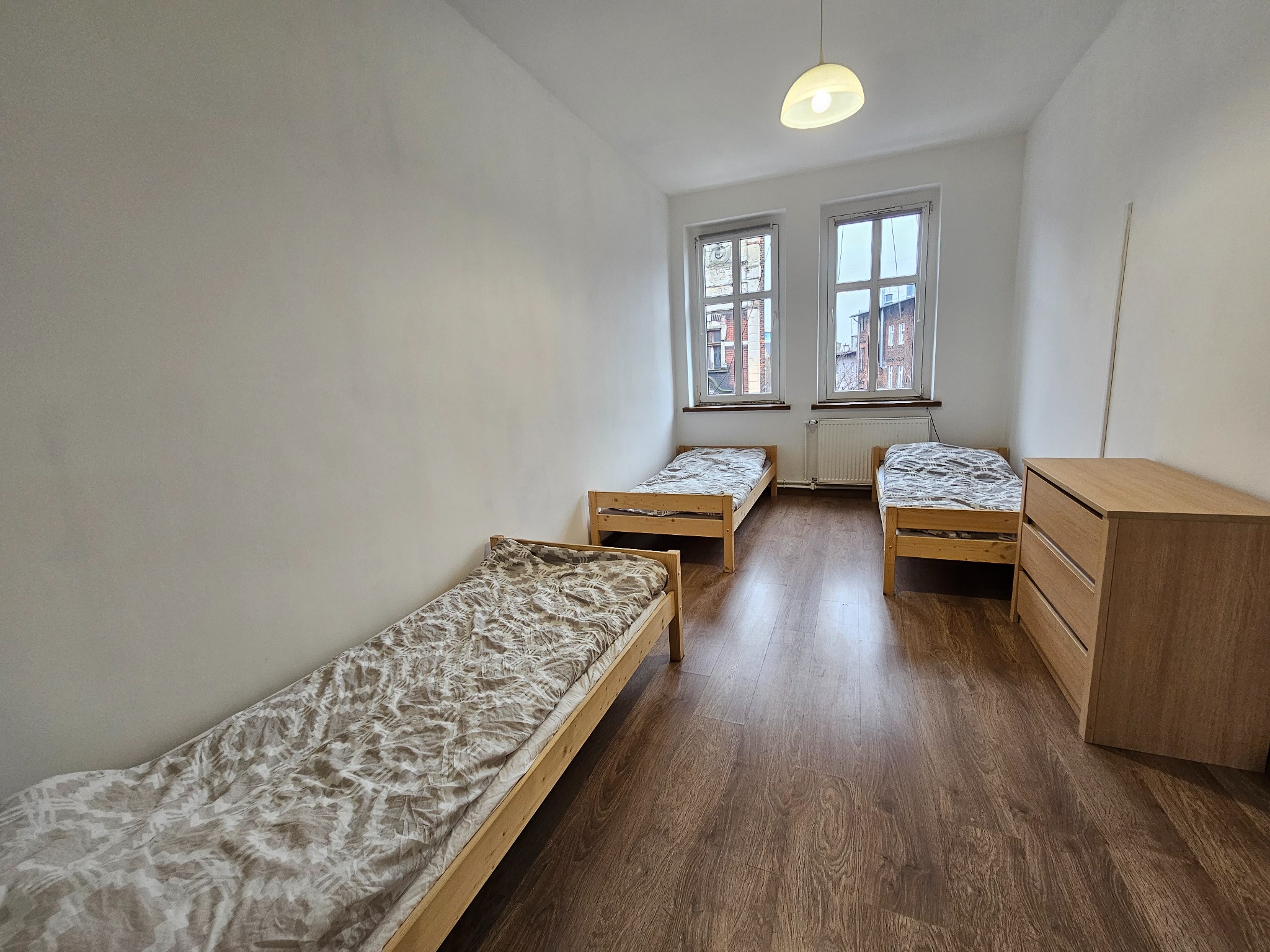 Pokój z trzema drewnianymi łóżkami pojedynczymi z pościelą w neutralnych kolorach, komodą i dwoma oknami z widokiem na budynki z czerwonej cegły.