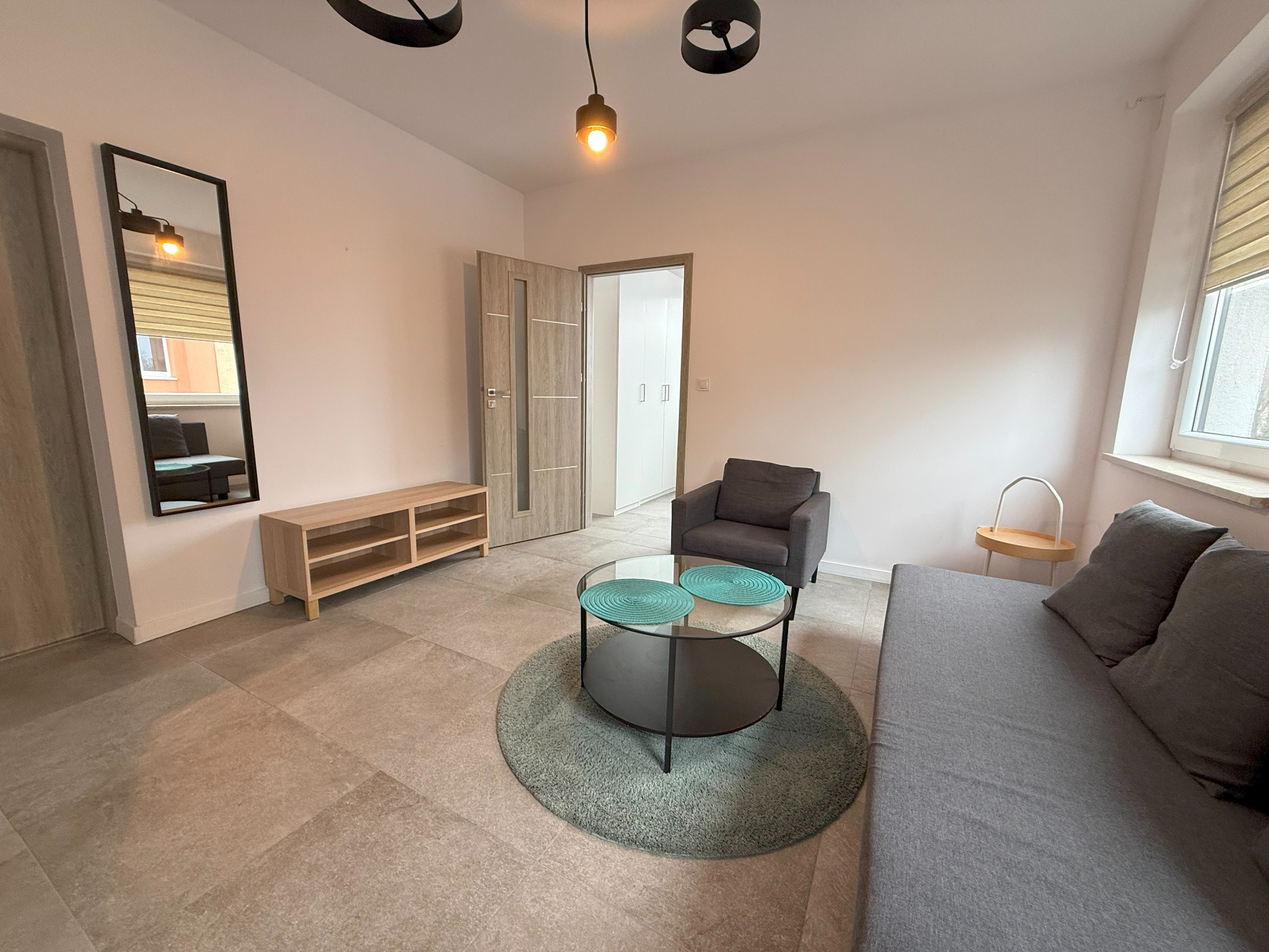 Jasne wnętrze pokoju z szarą sofą, fotelem, okrągłym stolikiem na puszystym dywanie i drewnianą szafką RTV, oświetlone trzema lampami wiszącymi.