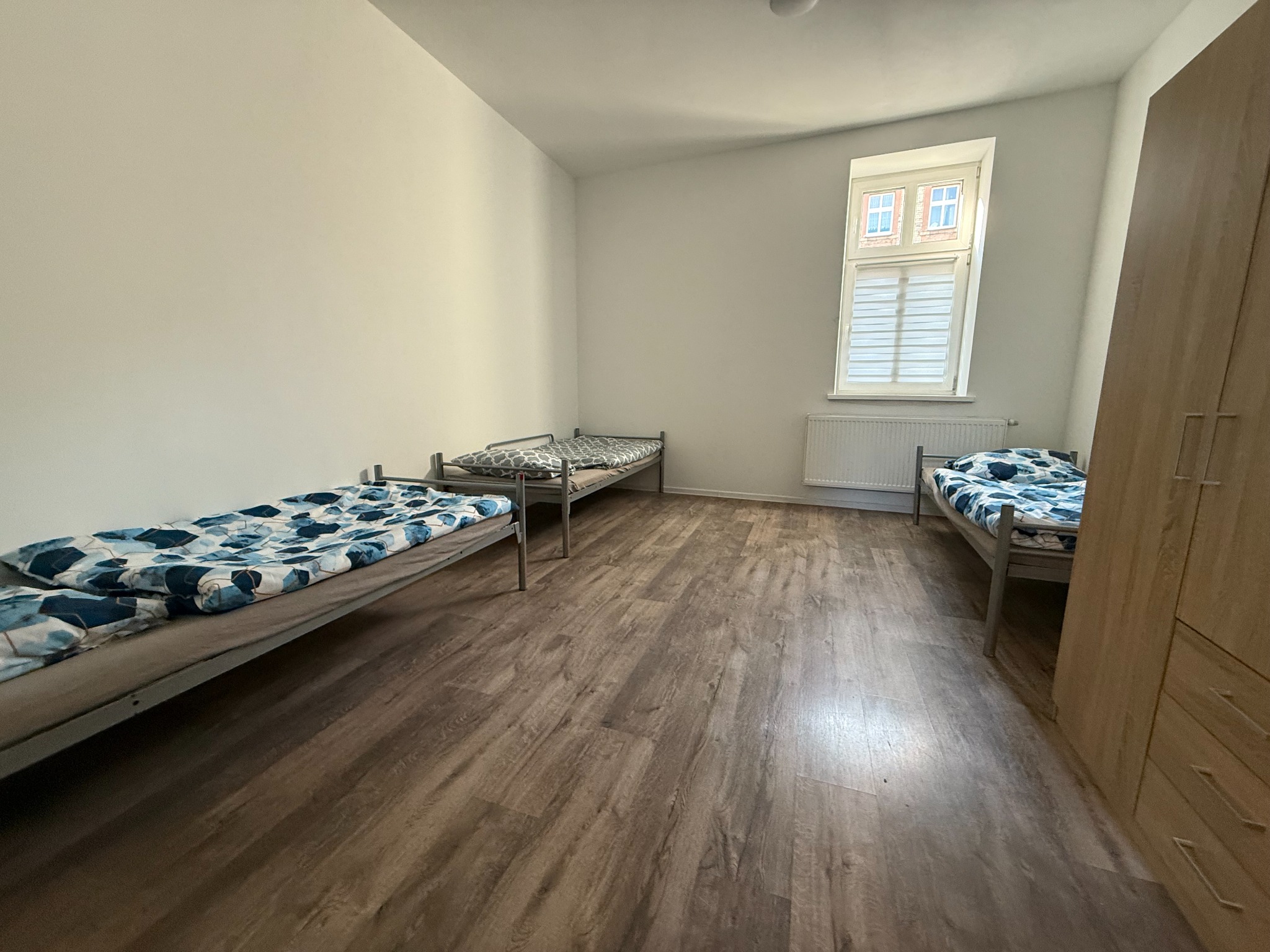 Prosty pokój z trzema łóżkami pojedynczymi, drewnianą podłogą i wysokim oknem z żaluzjami, ukazujący surowy, funkcjonalny styl.