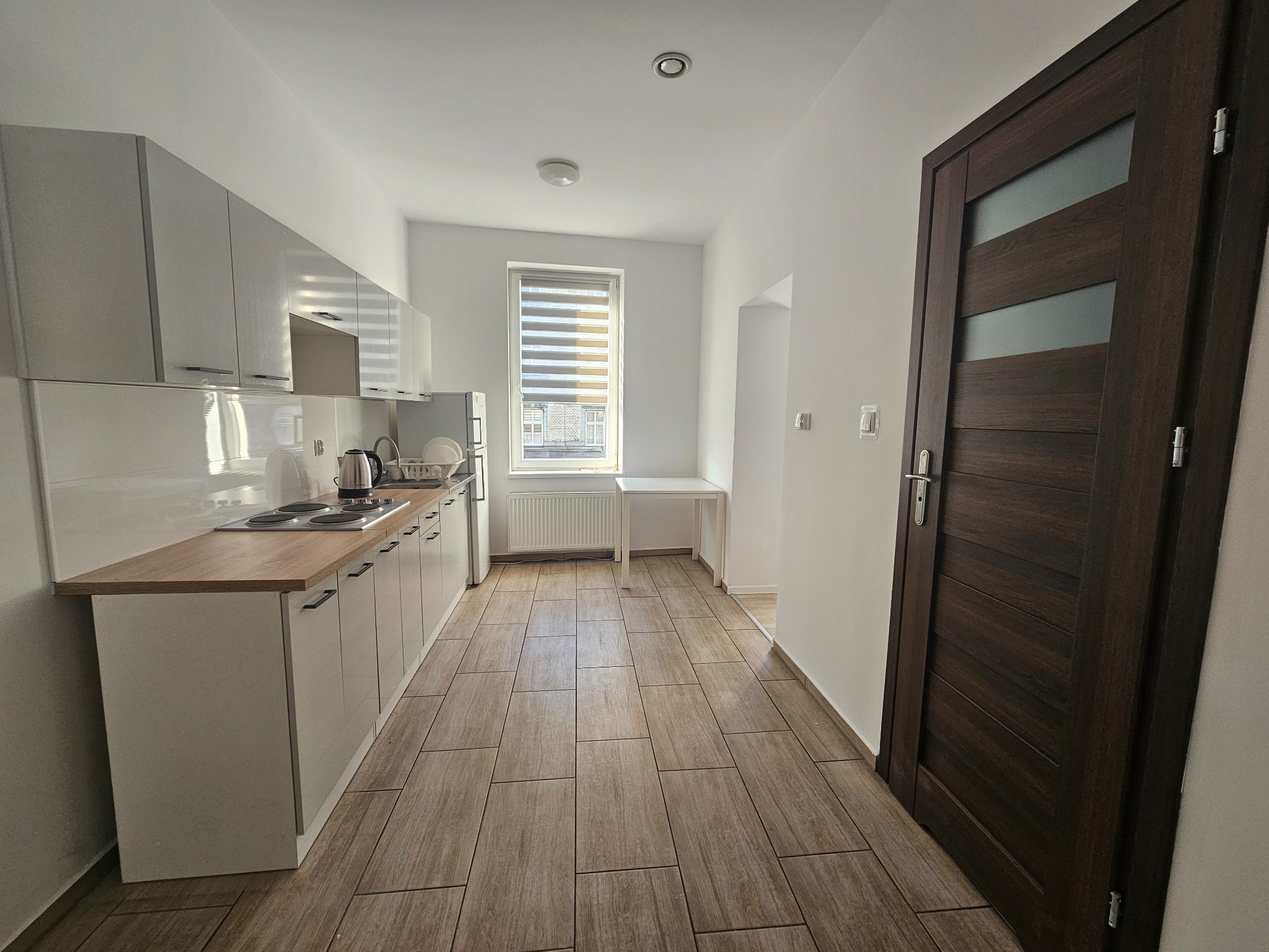 Wnętrze jasnej kuchni z białymi szafkami, drewnianym blatem, płytą grzewczą i czajnikiem elektrycznym, widok na okno z żaluzjami poziomymi oraz ciemne drzwi z przeszkleniami.