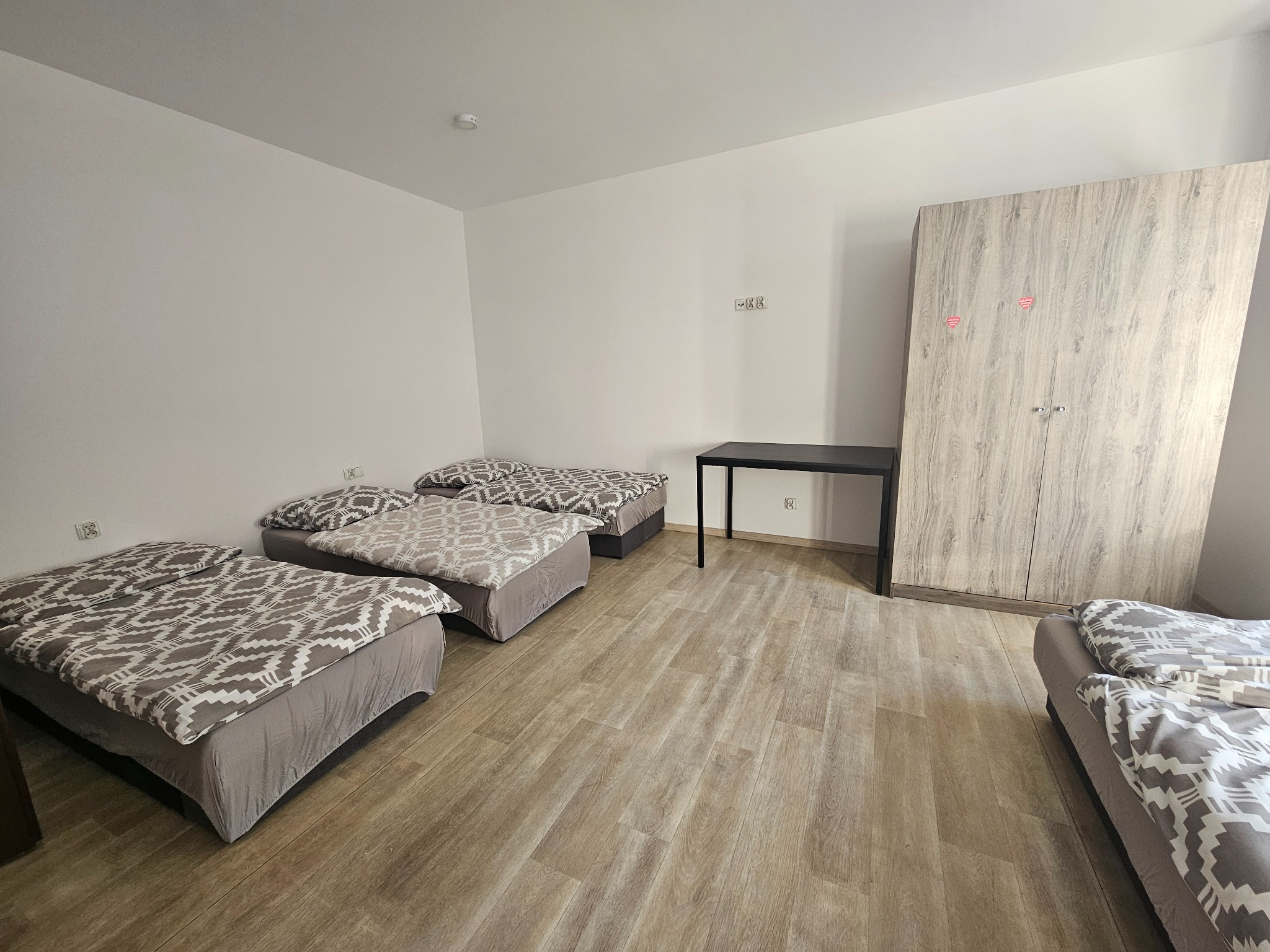 Wnętrze pokoju z czterema łóżkami pojedynczymi z szaro-białą pościelą w geometryczne wzory, czarnym biurkiem i dużą szafą z drewnianym wzorem na drzwiach.