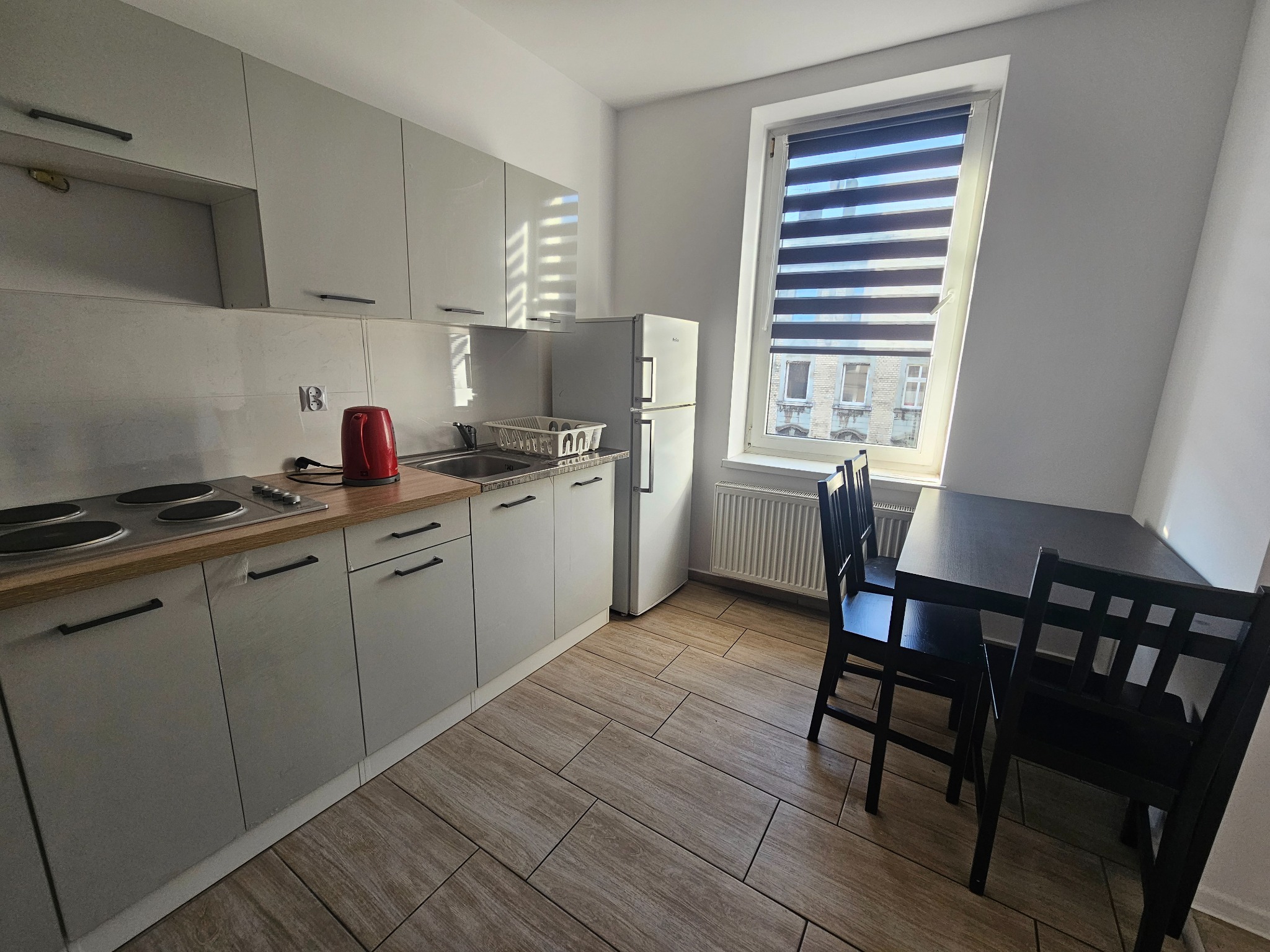 Jasna kuchnia z drewnianymi akcentami, stół z krzesłami przy oknie z żaluzjami poziomymi.