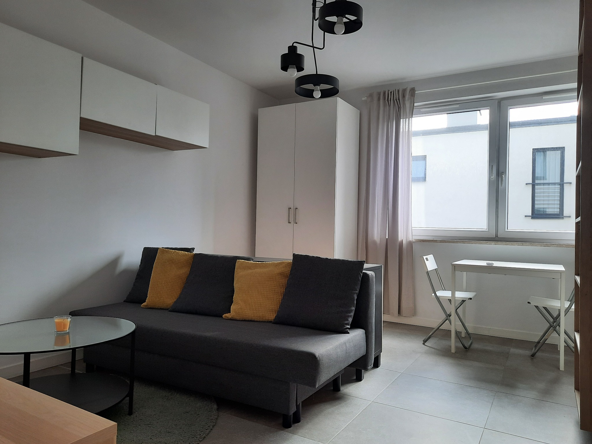 Jasne wnętrze pokoju z szarą rozkładaną sofą z żółtymi poduszkami, okrągłym stolikiem kawowym ze świecą, białymi szafkami wiszącymi i stołem z krzesłami pod oknem.