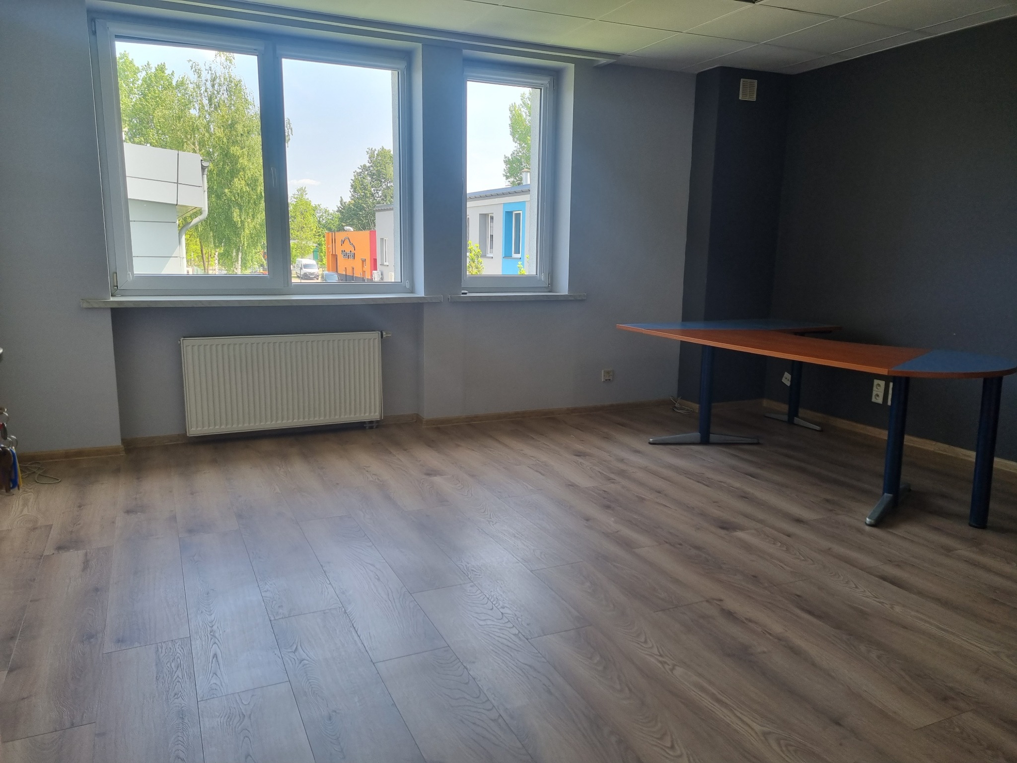 Puste biuro z dwoma oknami, grzejnikiem i stołem z blatem w kolorze drewna i niebieskimi krawędziami, z widokiem na zewnątrz.
