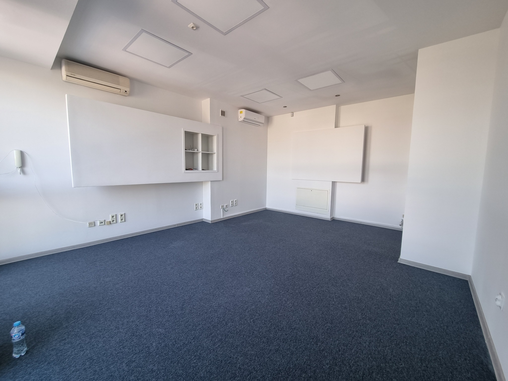 Puste biuro z szarą wykładziną, białymi ścianami, klimatyzacją i butelką wody w rogu.
