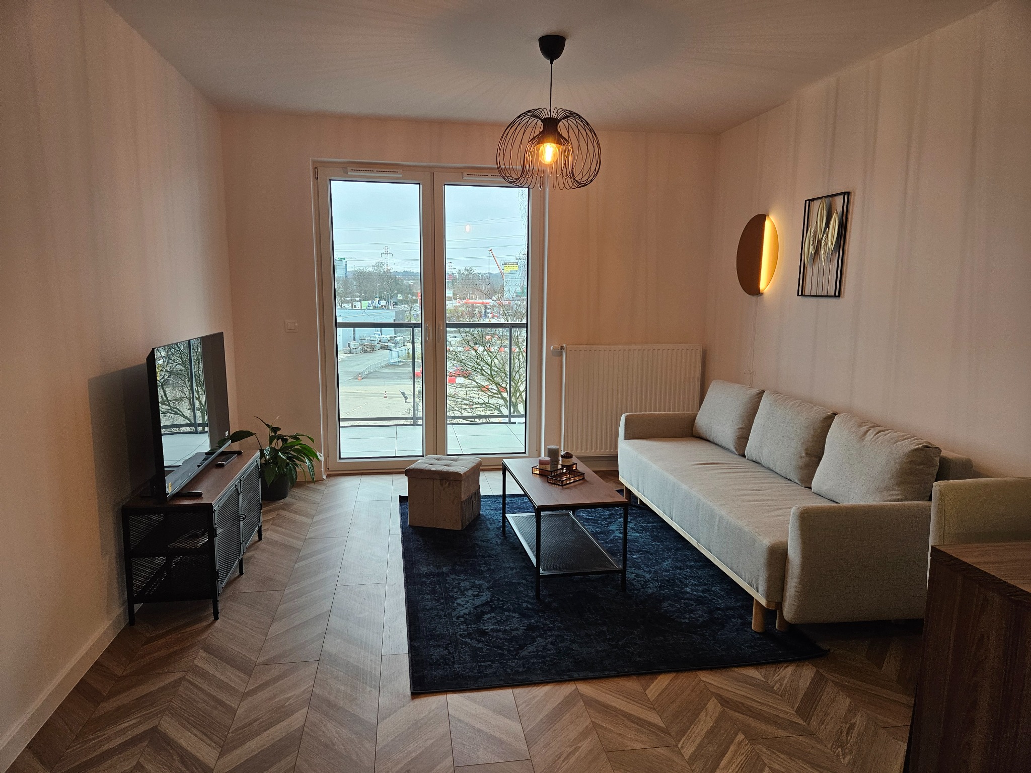 Jasne, nowocześnie urządzone mieszkanie z parkietem w jodełkę, szarą sofą i czarnym dywanem. Widok z okna na plac budowy.