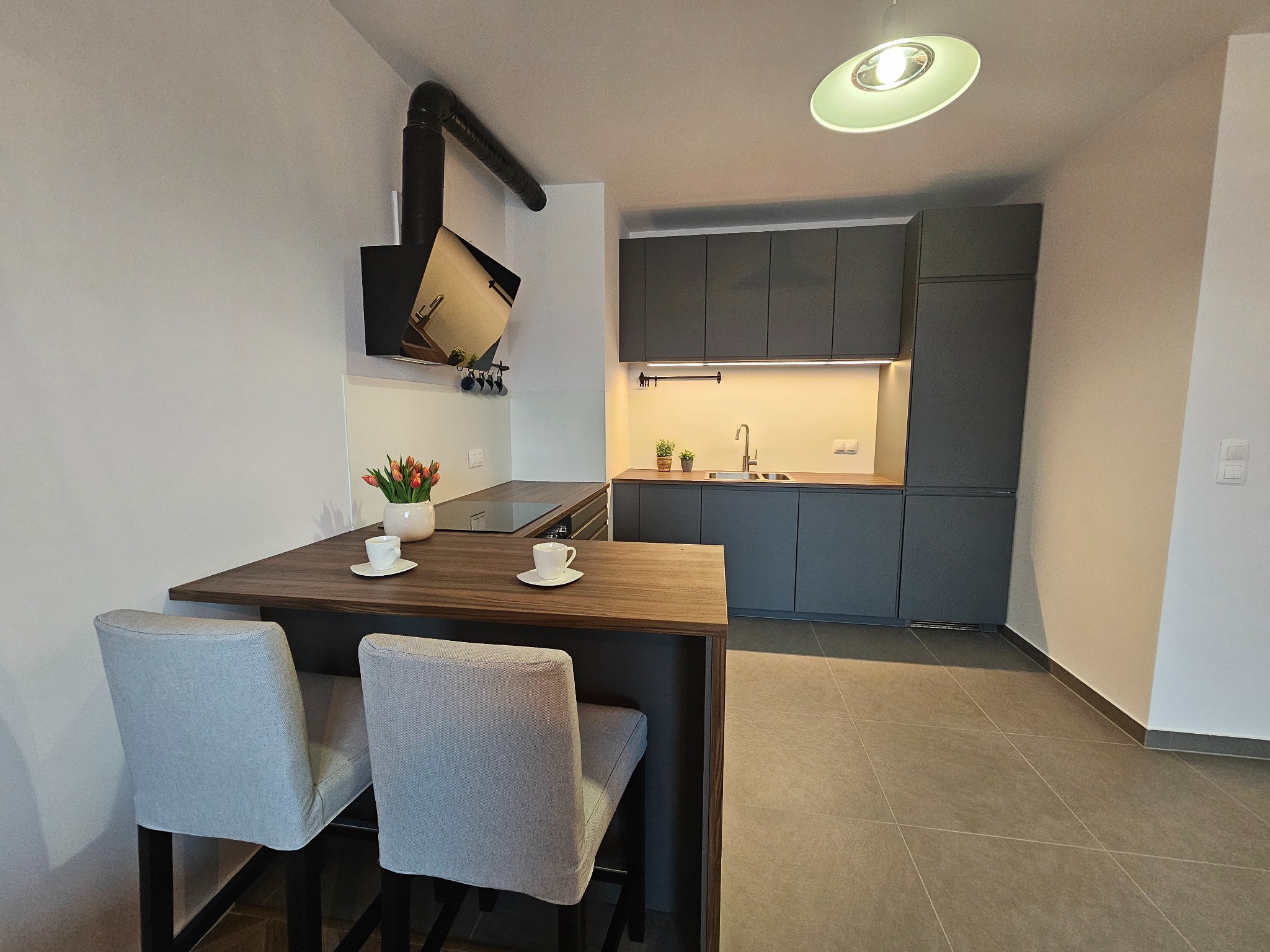 Nowoczesna kuchnia z szarymi szafkami, drewnianym blatem, wyspą z dwoma krzesłami barowymi, kwiatami w wazonie i filiżankami na blacie.