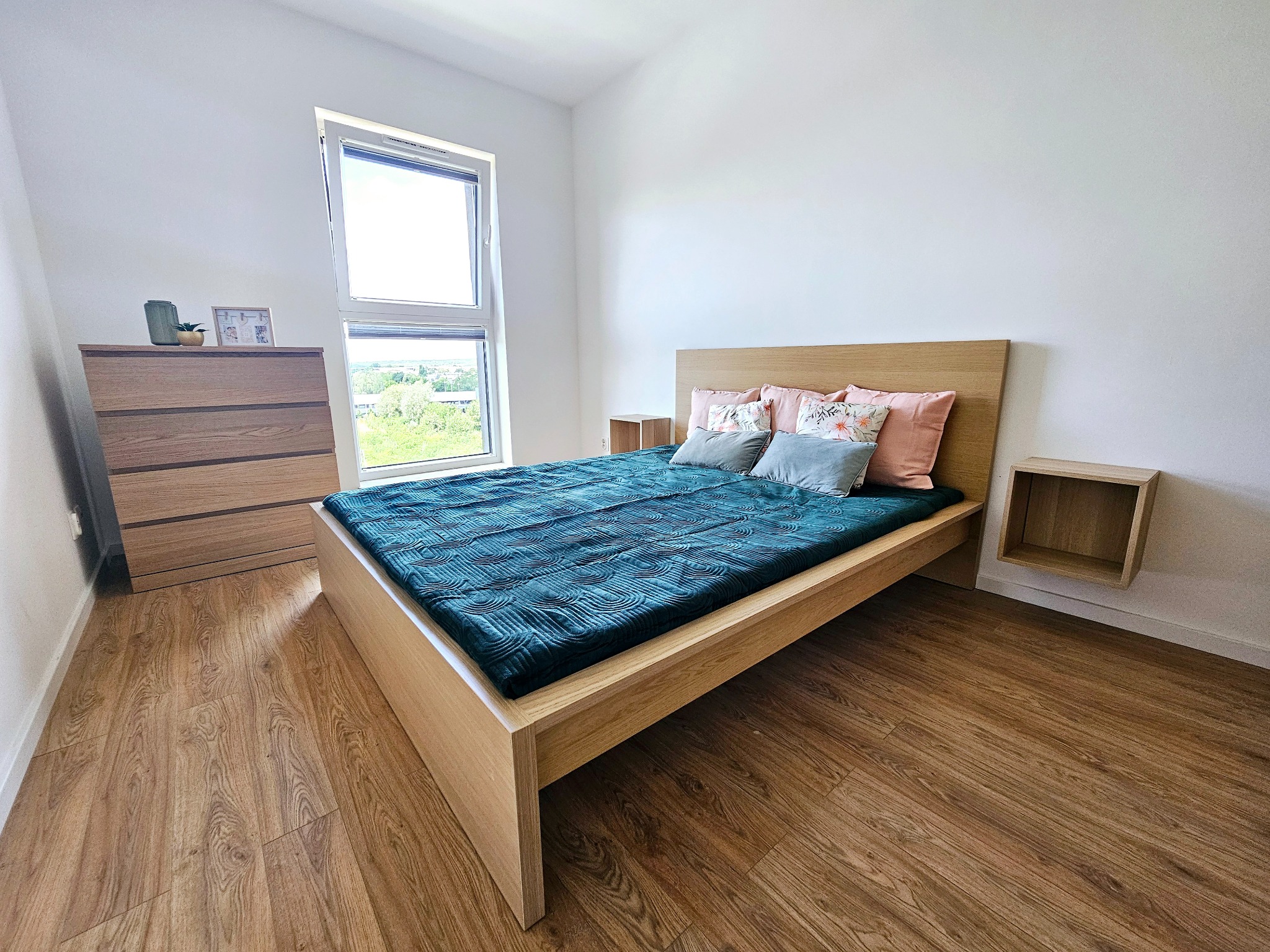 Jasne wnętrze sypialni z drewnianym łóżkiem, turkusową narzutą i widokiem z okna na zieleń.