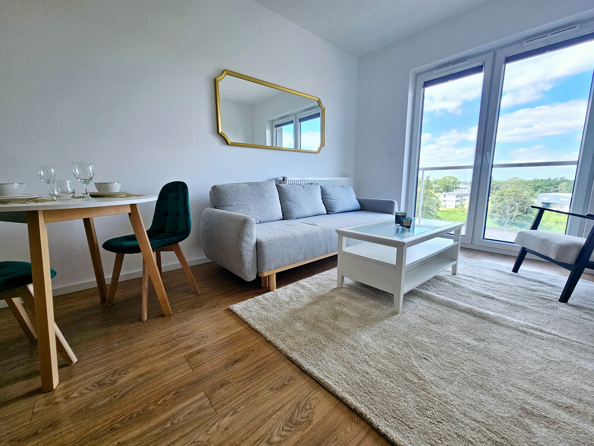 Jasne wnętrze pokoju z drewnianą podłogą, szarą sofą z poduszkami, białym stolikiem kawowym, okrągłym stołem z nakryciem i zielonymi krzesłami, lustrem w złotej ramie i dużym oknem z widokiem...