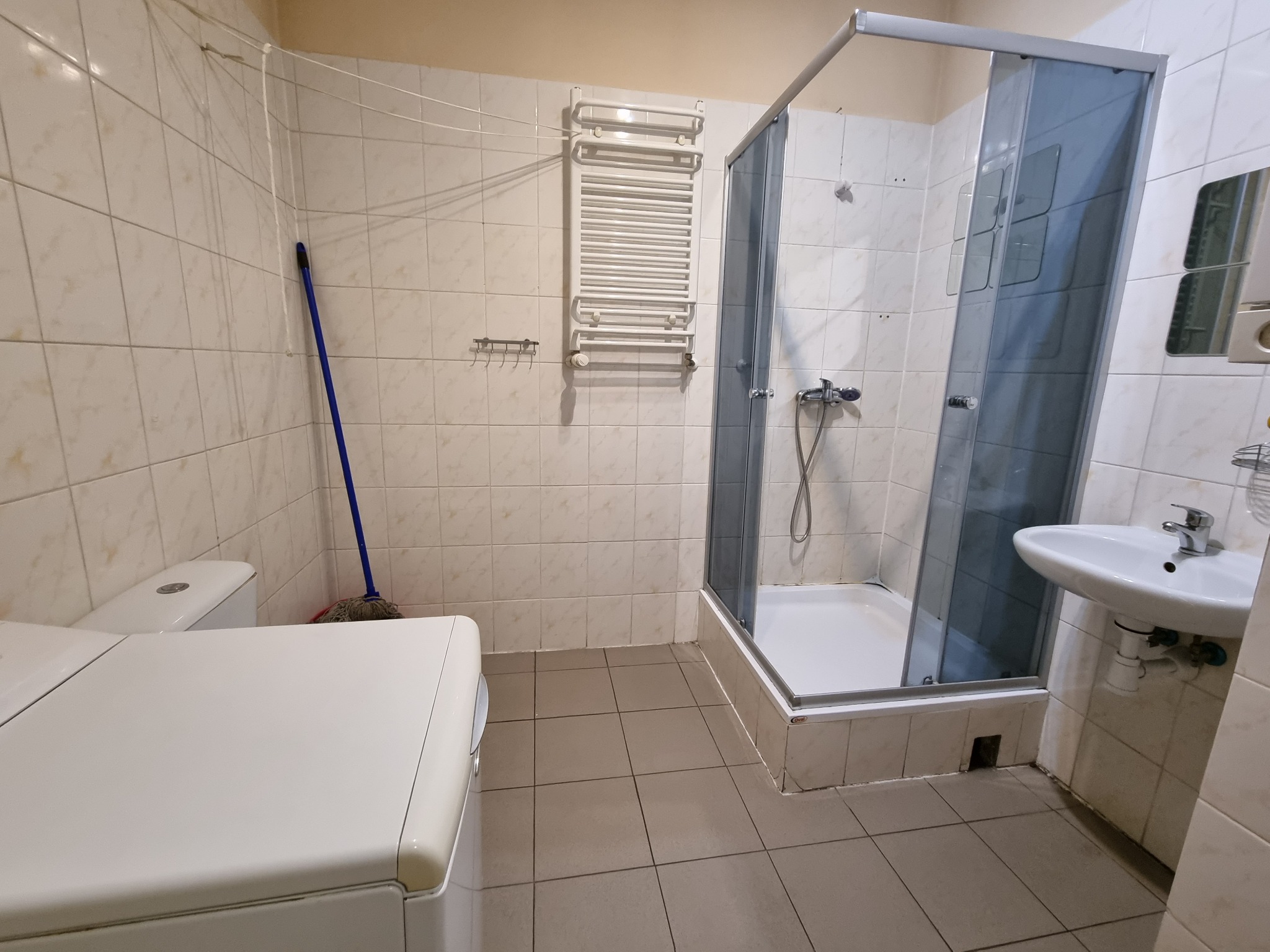 Wnętrze łazienki z kabiną prysznicową, pralką i grzejnikiem drabinkowym, widoczne płytki w beżowym odcieniu i mop oparty o ścianę.