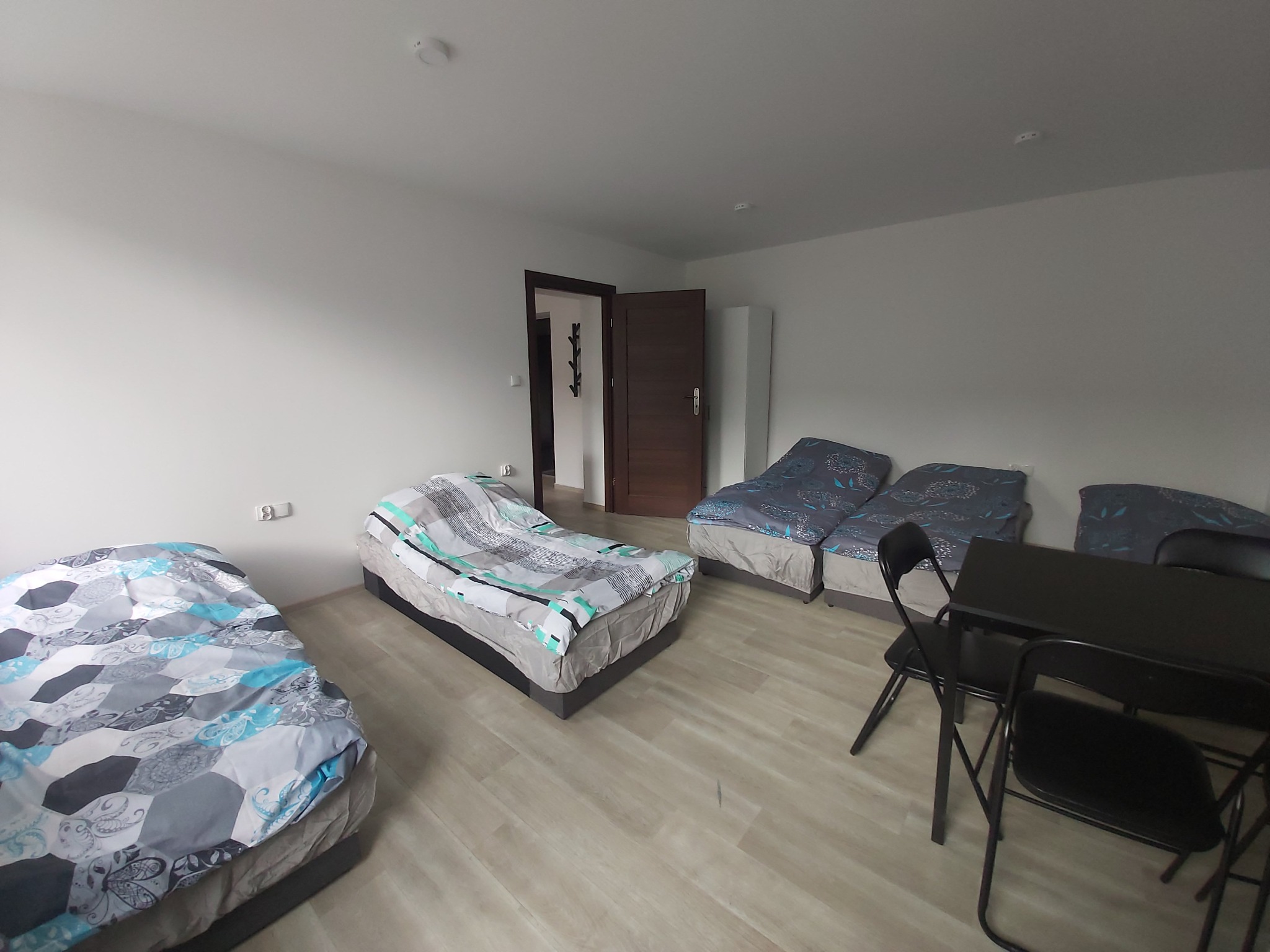 Pokój z trzema łóżkami pojedynczymi z pościelą w geometryczne wzory, czarnym stołem i krzesłami, widok na otwarte drzwi i białą szafę w tle.