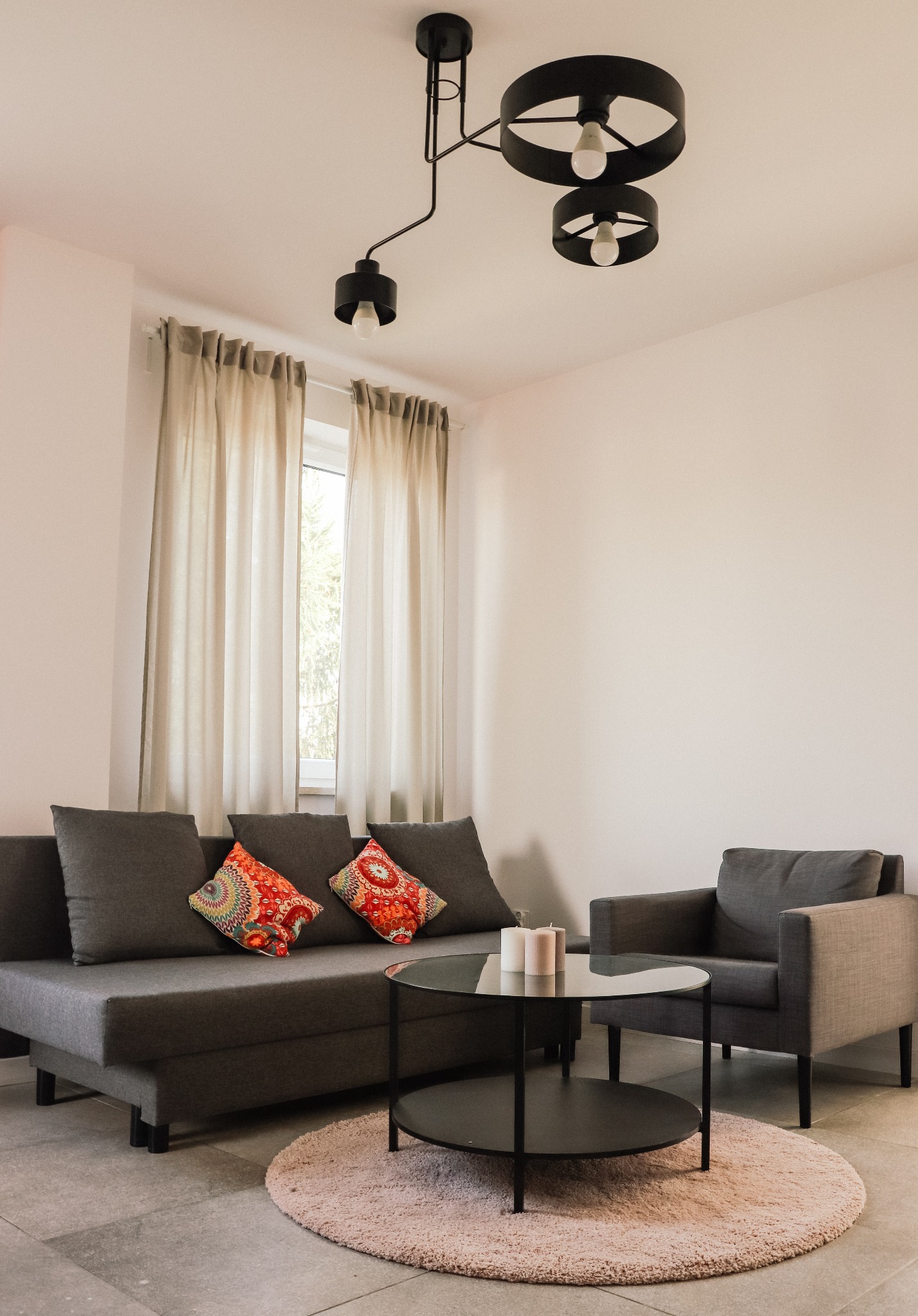 Nowoczesny salon z szarą sofą z kolorowymi poduszkami, okrągłym stolikiem kawowym z czarnym blatem, szarym fotelem i designerską lampą sufitową.