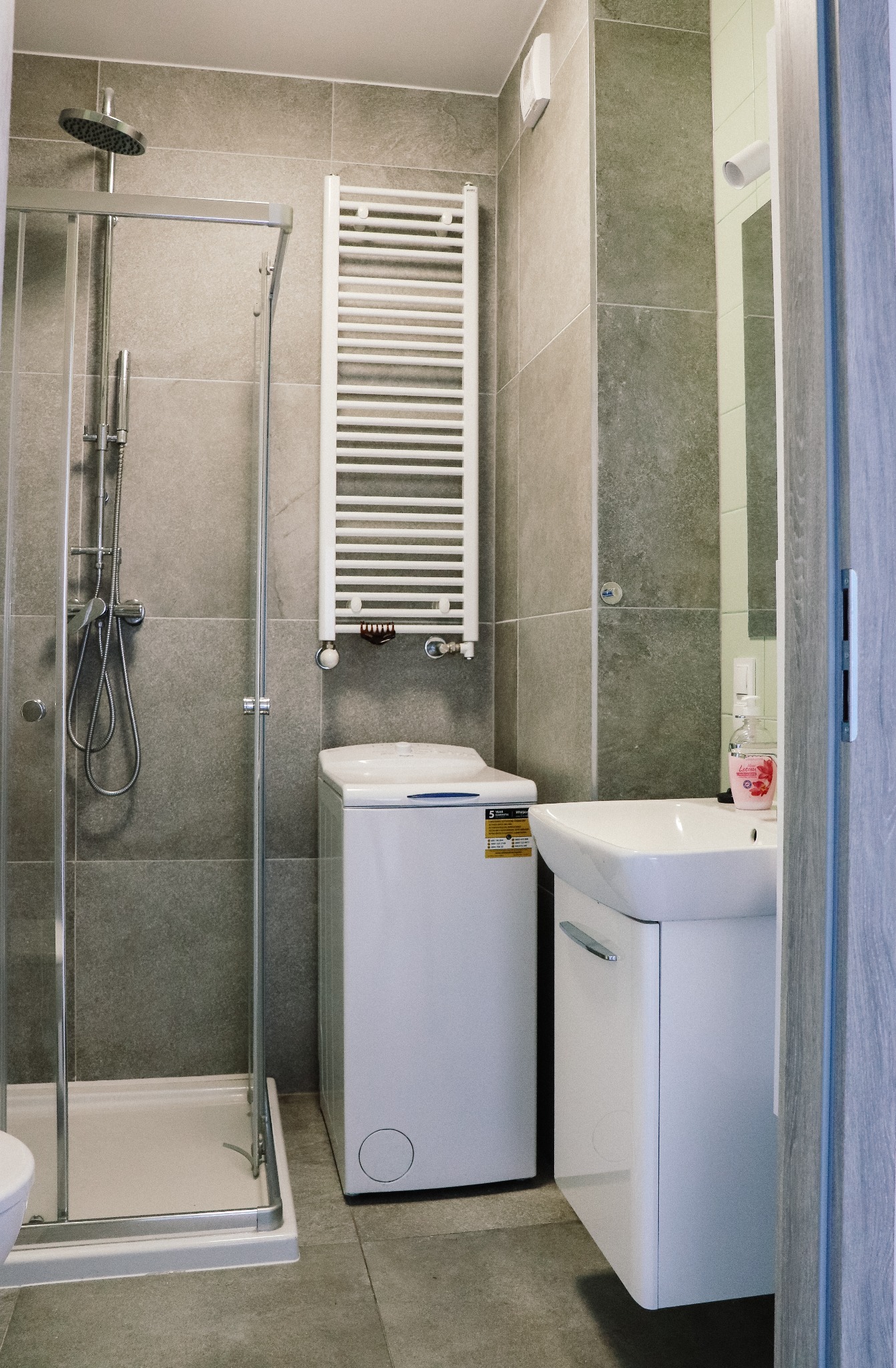 Wnętrze łazienki w odcieniach szarości z kabiną prysznicową, pralką i umywalką z szafką, widoczne z perspektywy drzwi.