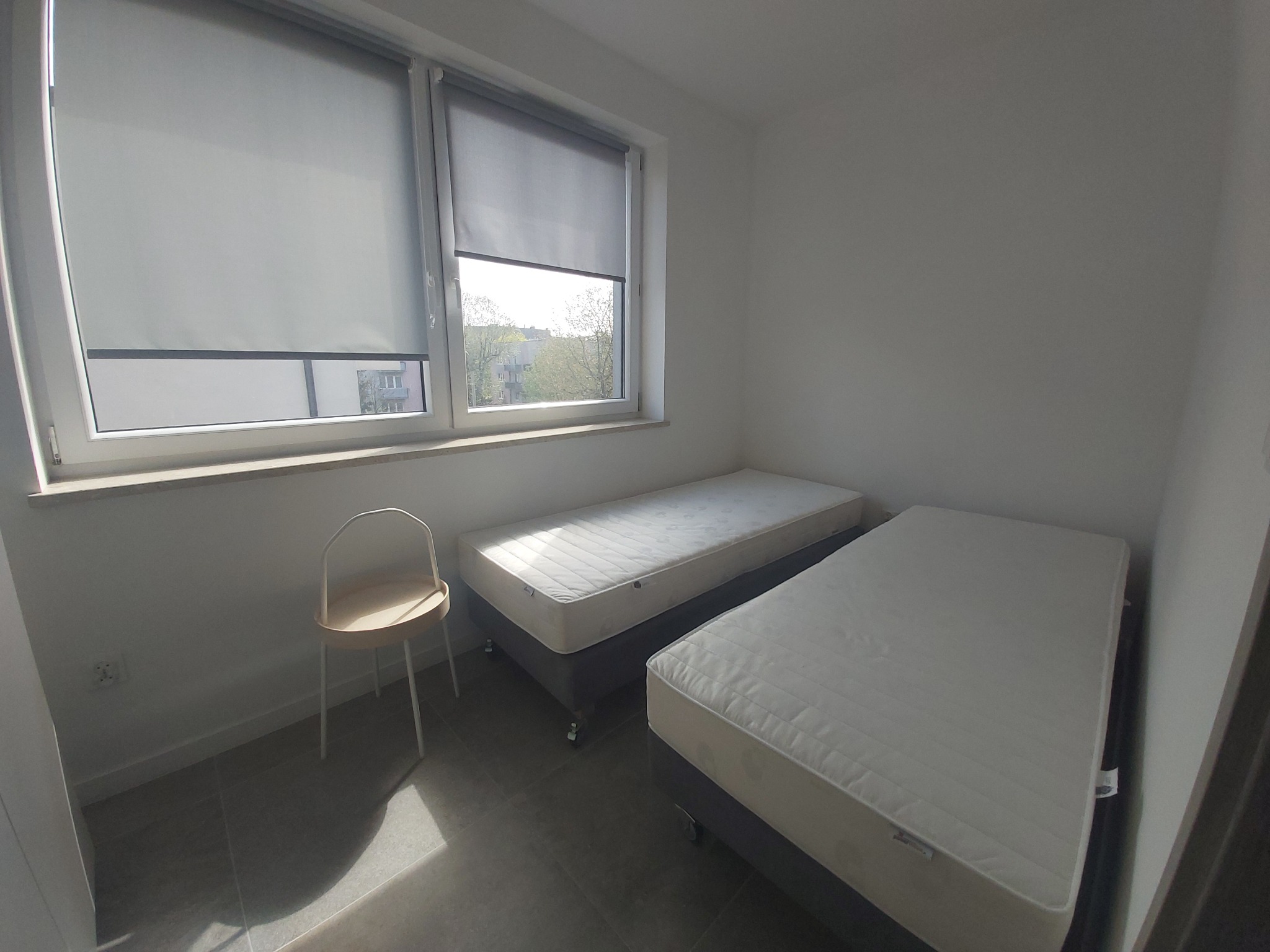 Pokój z dwoma pojedynczymi łóżkami, krzesłem i zasłoniętymi roletami w oknach, widok na budynek za oknem.