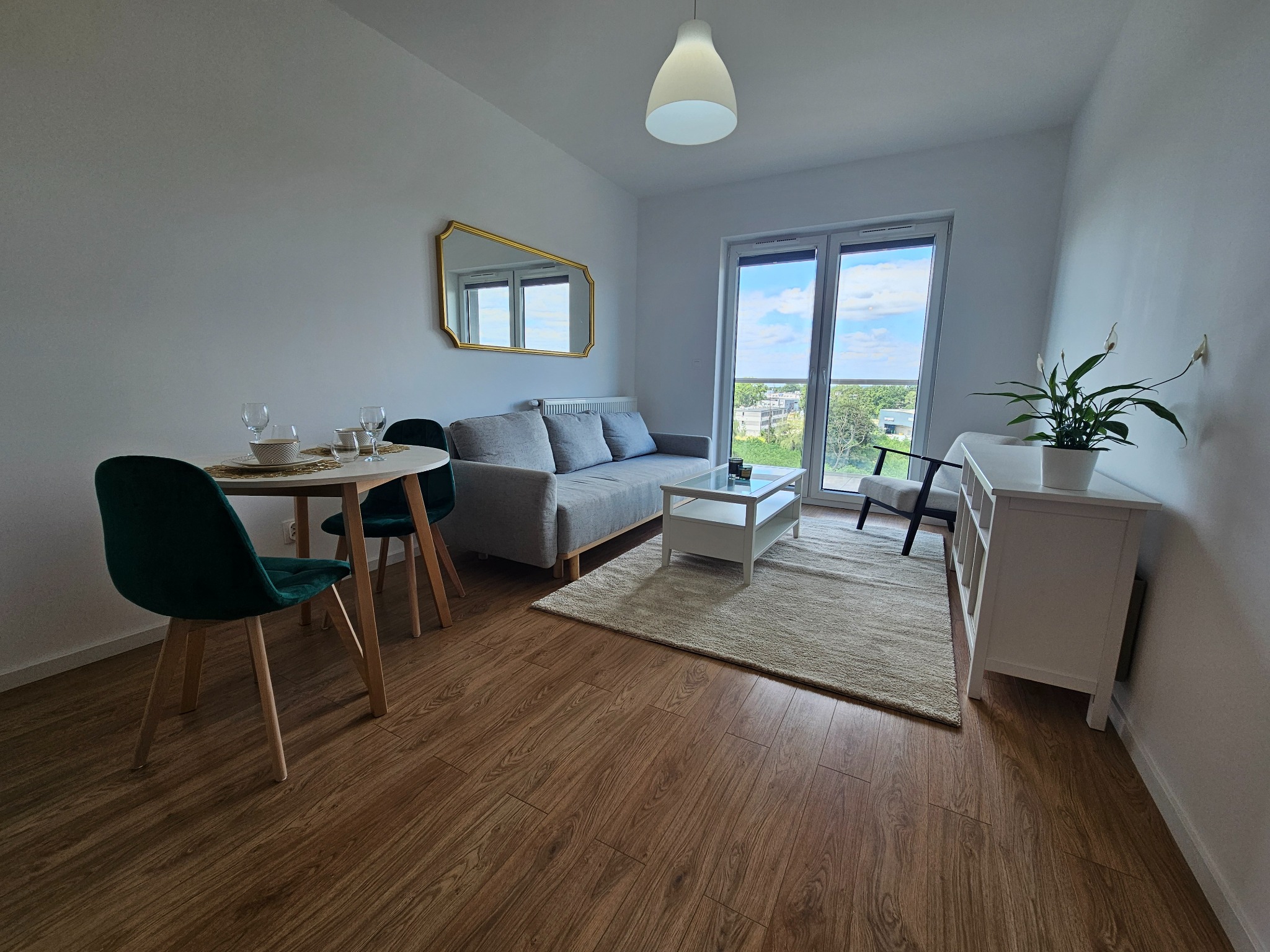 Jasne wnętrze mieszkania z drewnianą podłogą, szarą sofą, okrągłym stołem z zielonymi krzesłami i lustrem w złotej ramie.