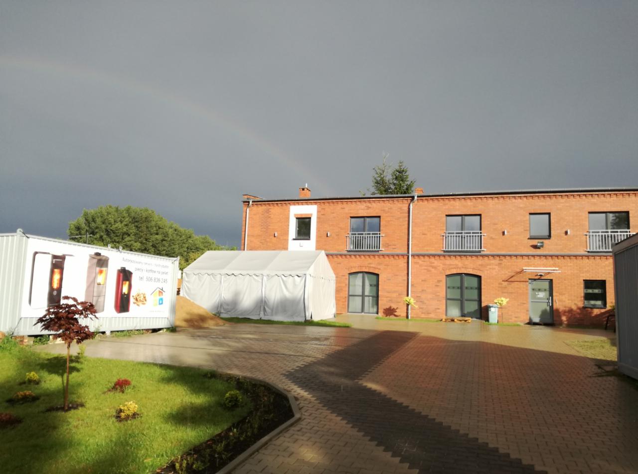 Ekspozycja zewnętrzna z kontenerem prezentującym modele pieców na paliwo stałe, obok namiot i budynek z czerwonej cegły pod pochmurnym niebem z fragmentem tęczy.